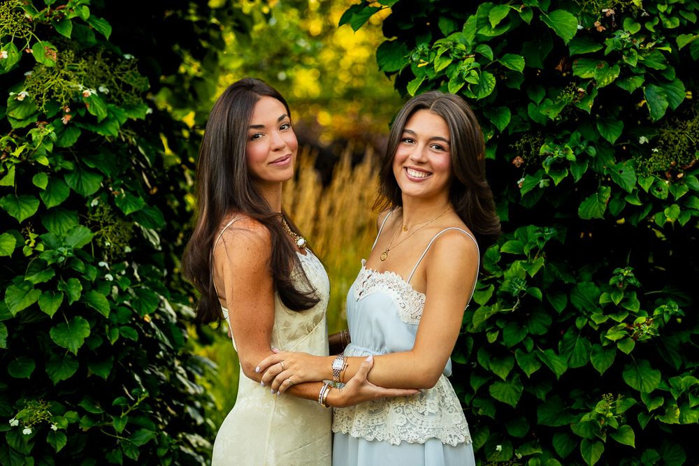 Two women in formal dresses pose together in a garden setting surrounded by lush green foliage.