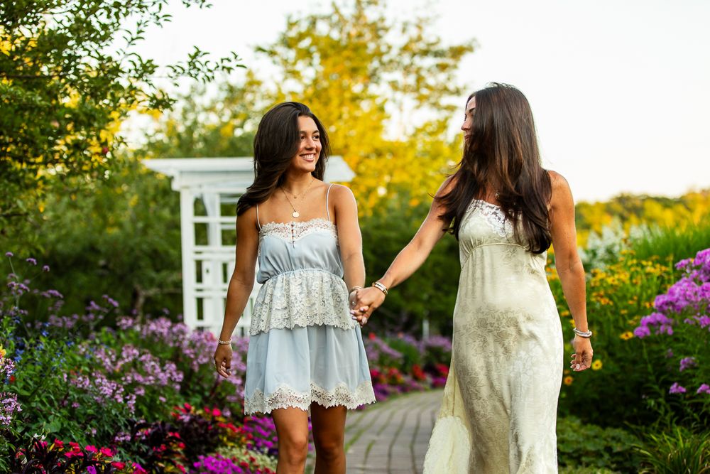 Two friends in summer dresses walk hand in hand through a blooming garden path at sunset.