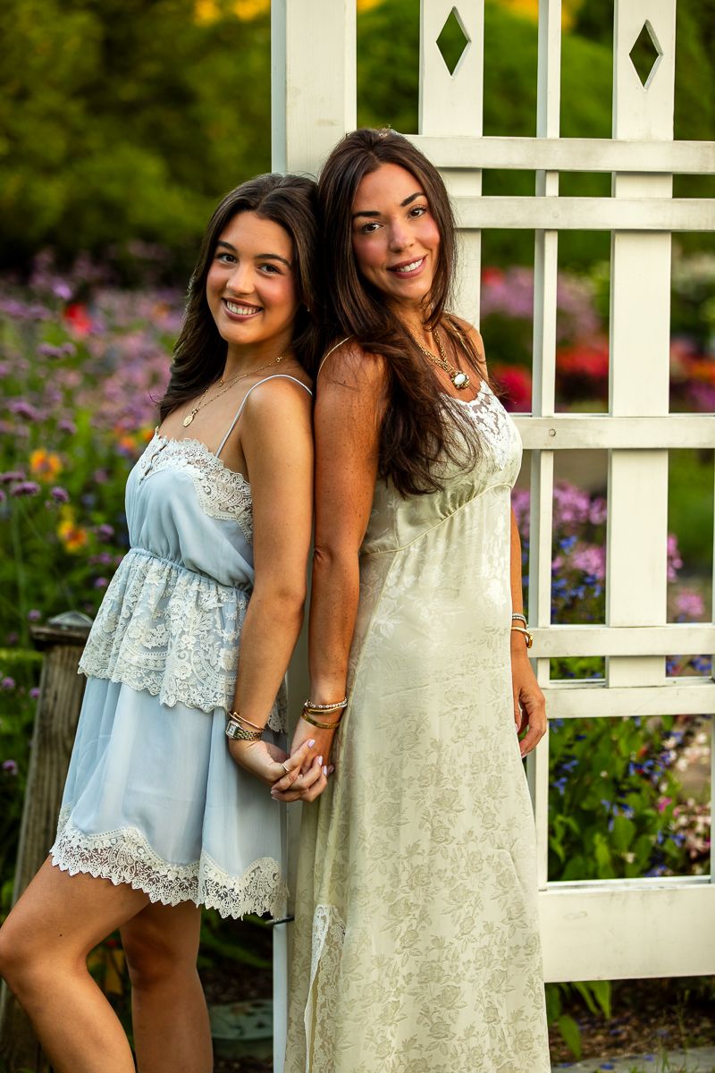 Two friends in light-colored dresses pose together by a white garden trellis surrounded by flowers.