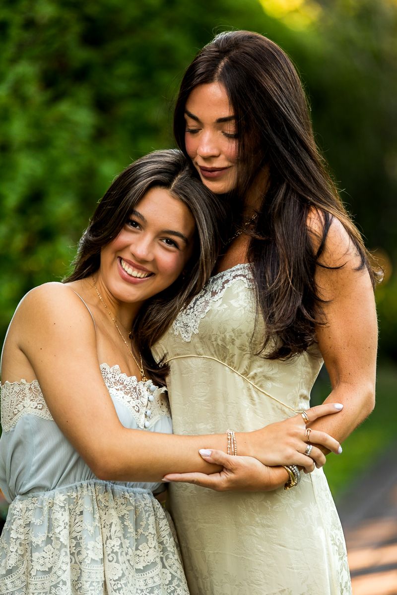 Two friends embracing and smiling while wearing elegant lace dresses in an outdoor garden setting.