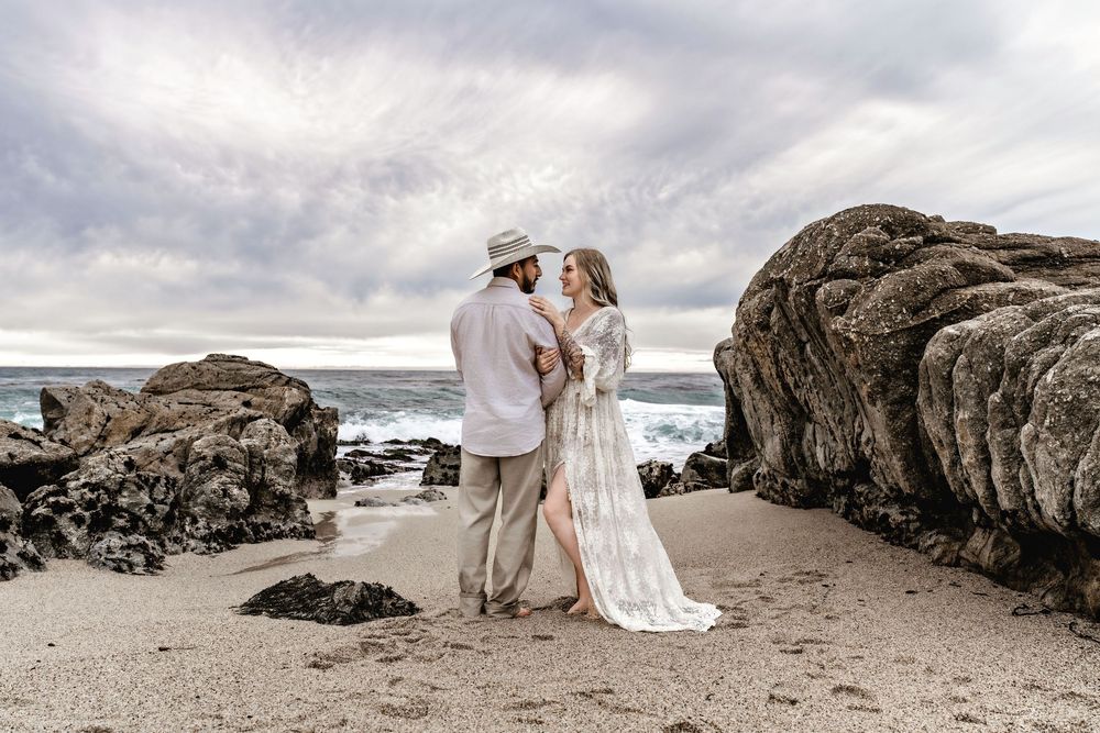 A romantic beach wedding photoshoot with rocks and dramatic cloudy skies in the background.