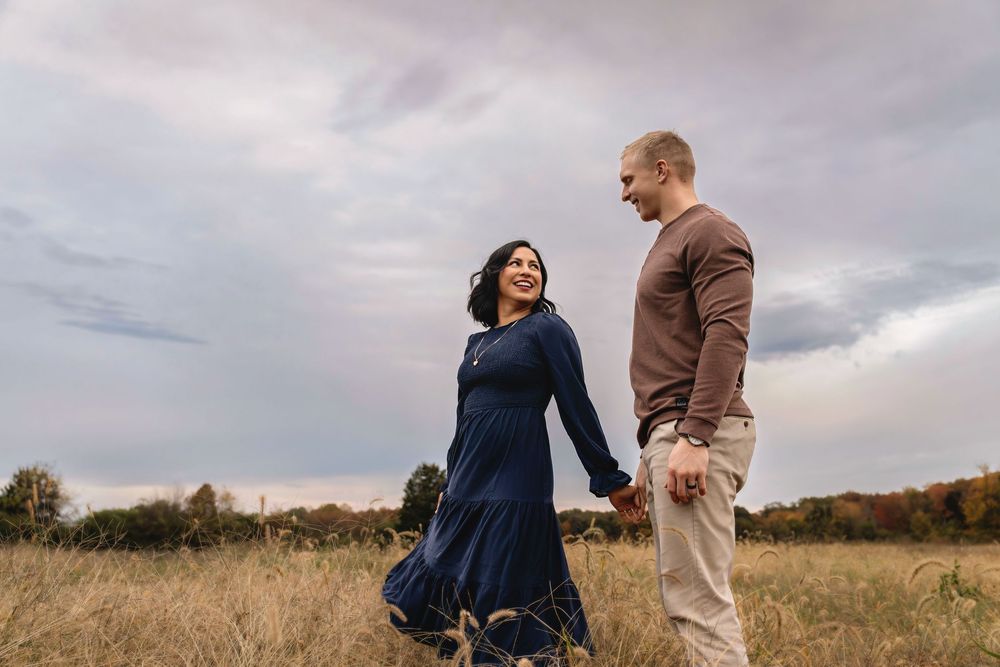 Couple shares romantic moments in an autumn field with moody cloudy sky at sunset.
