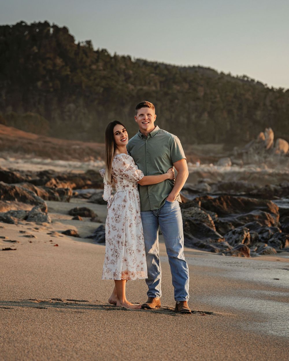 A couple strolls along a scenic coastal beach with rocky cliffs in the background at sunset.