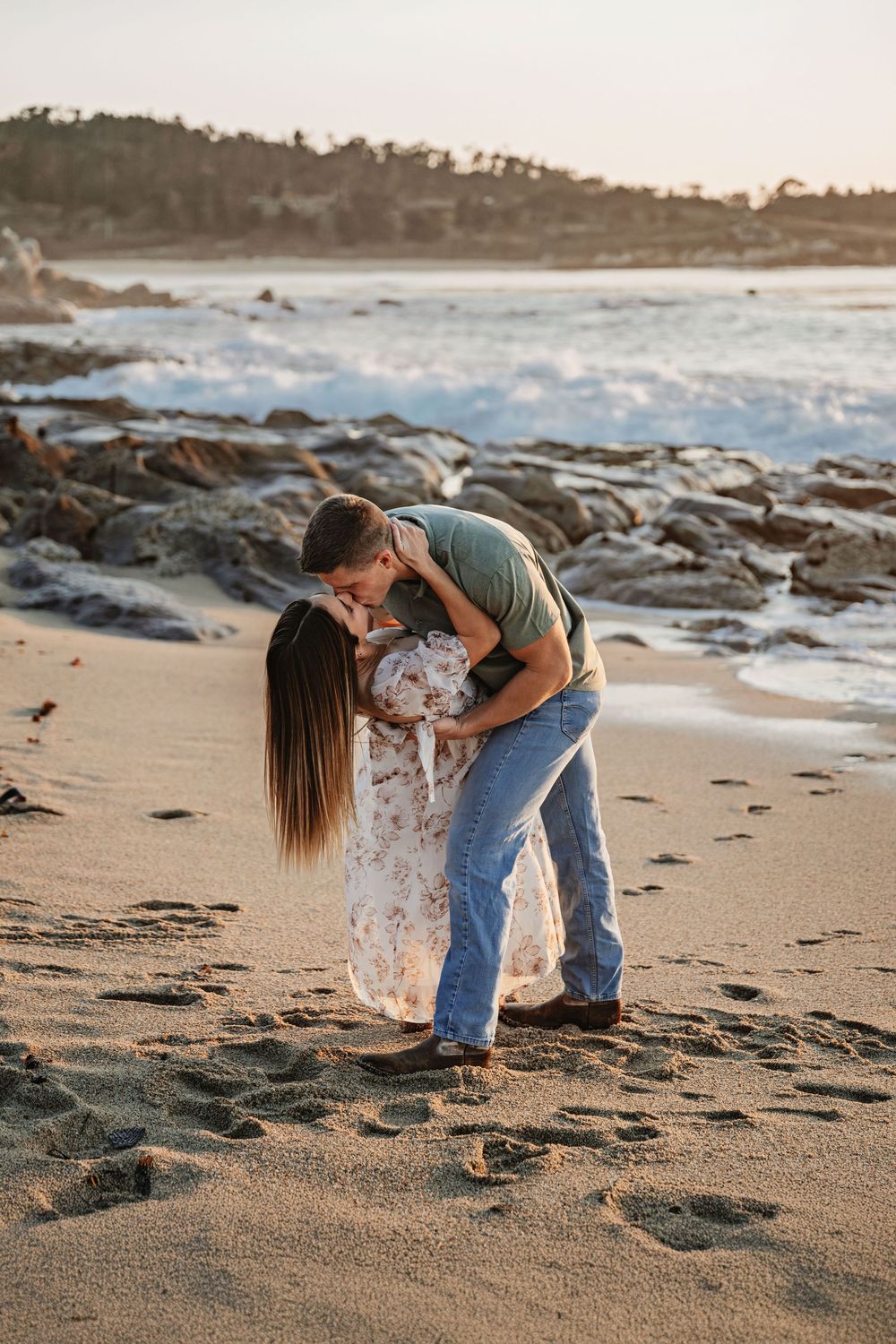 Couple shares tender moment on sandy beach with crashing waves and rocky coastline at sunset.