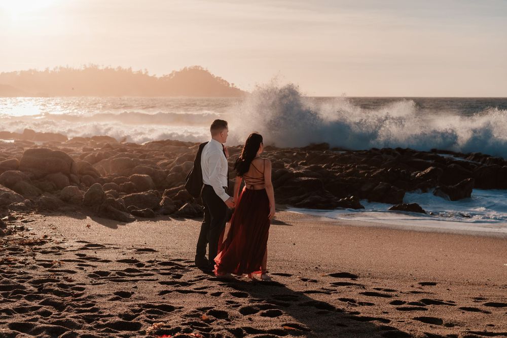 Dramatic ocean waves crash against rocks as silhouettes stand on beach at sunset.