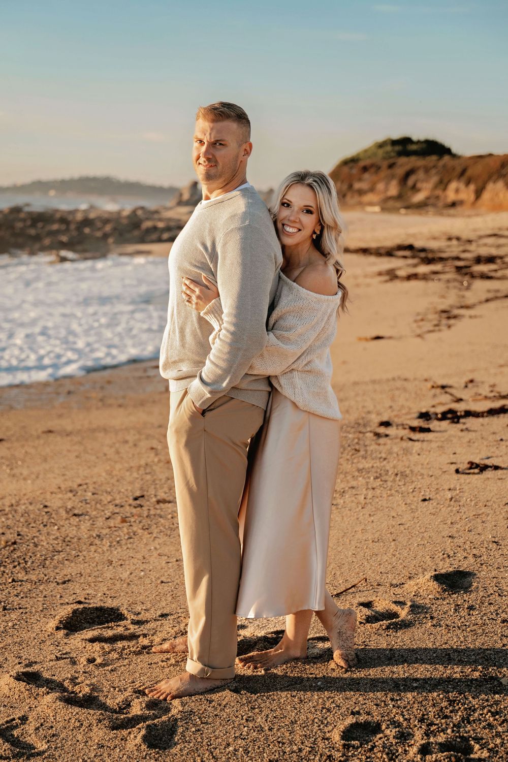 Couple in neutral cream outfits embrace while walking along a scenic coastal beach at sunset.