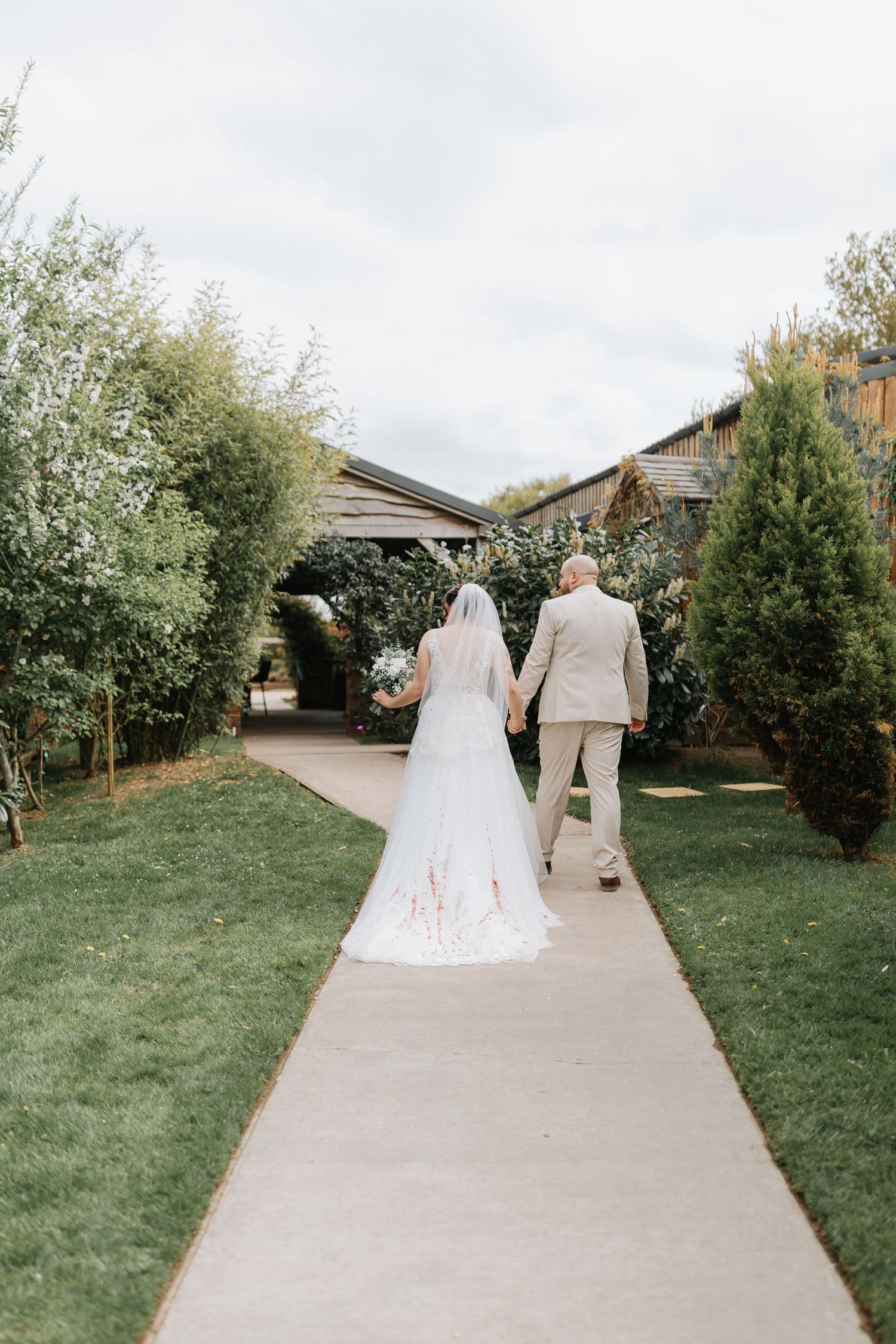 A couple in wedding attire walks down a concrete path surrounded by trees and grass in a garden setting.