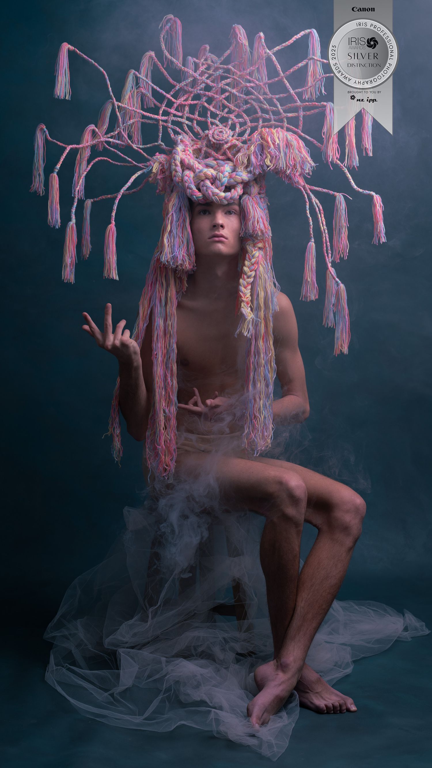 A man wears an elaborate headpiece made out of wool crochet and wire. He sits in an awkward pose that looks like a tree.