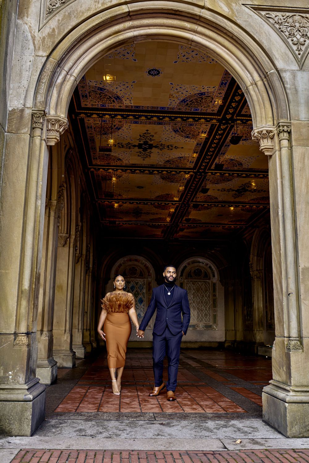 Two people walk through an ornate archway in Central Park's Bethesda Terrace tunnel with decorative ceiling tiles. Engagement photoshoot