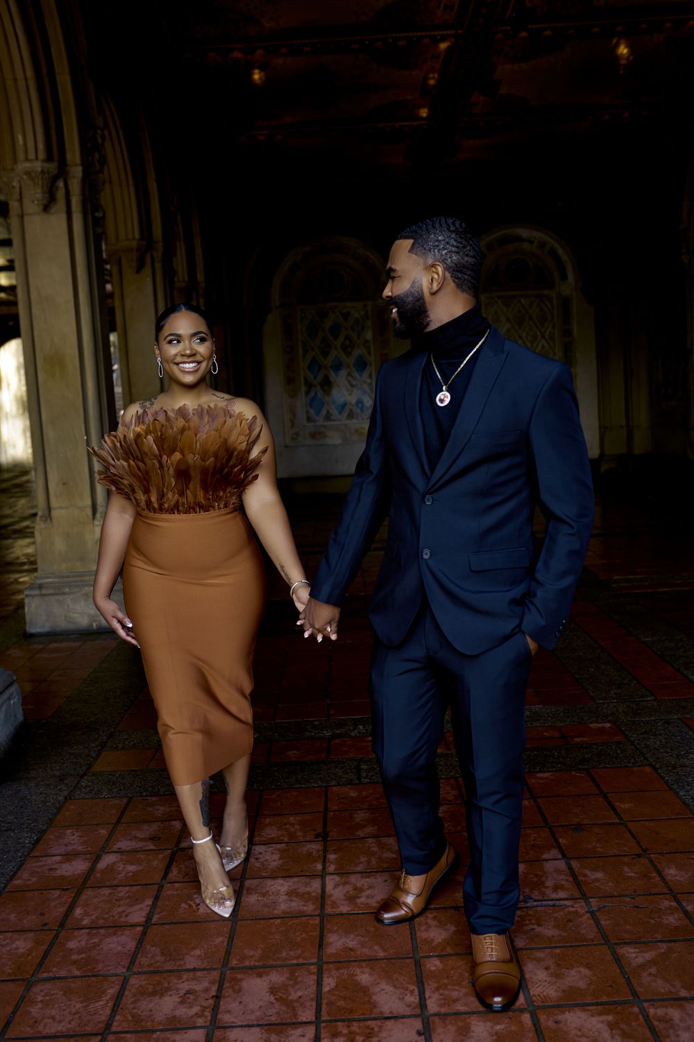 A stylish couple holds hands while walking through an elegant corridor, wearing formal brown dress and navy suit. Engagement photoshoot