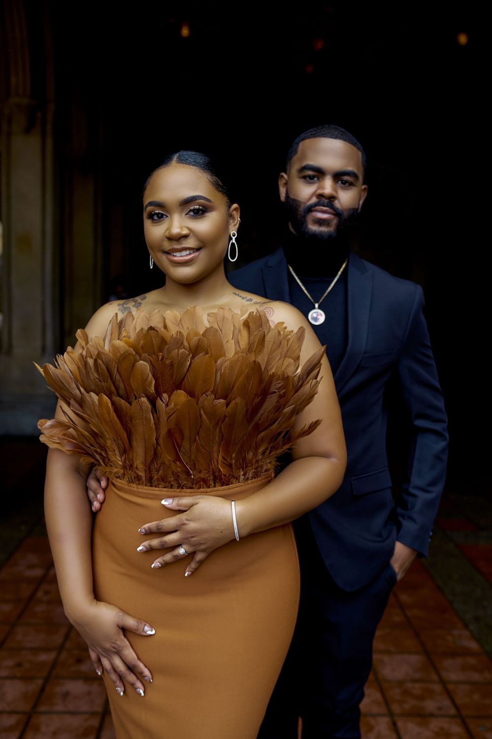 A couple poses elegantly together, with a brown feathered dress design and navy blue suit against a dim background. Engagement photoshoot