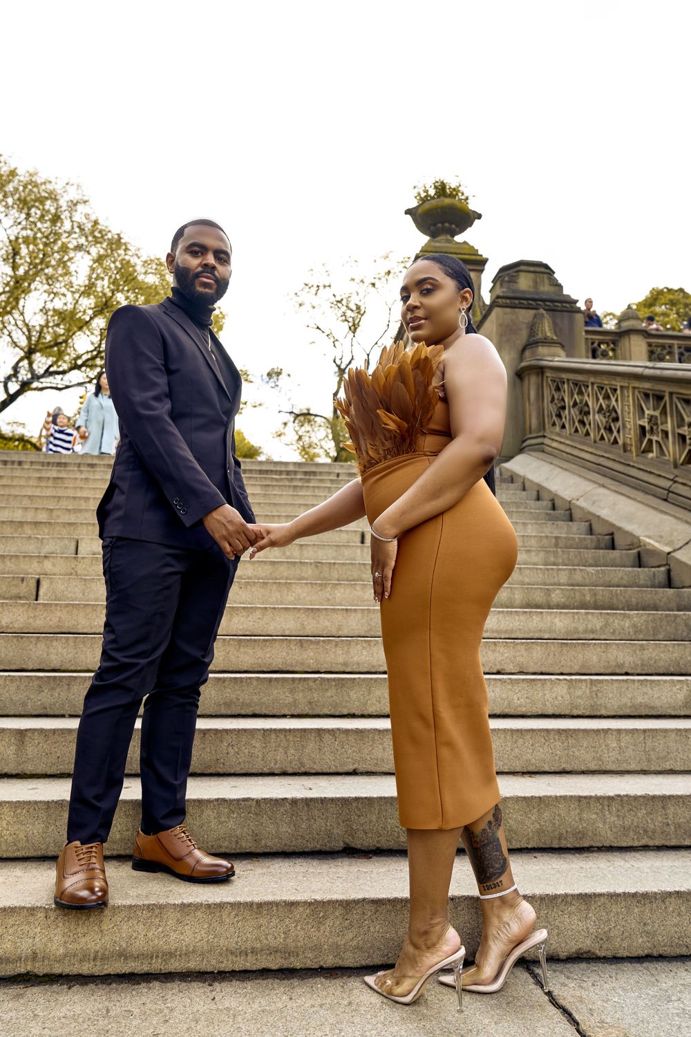 A couple in formal wear dances playfully on stone steps outside, with him in navy and her in camel-colored dress.