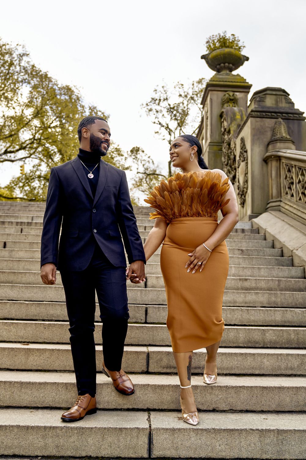 A couple holds hands while standing on stone steps, wearing elegant formal attire in navy blue and caramel colors. Engagement photoshoot