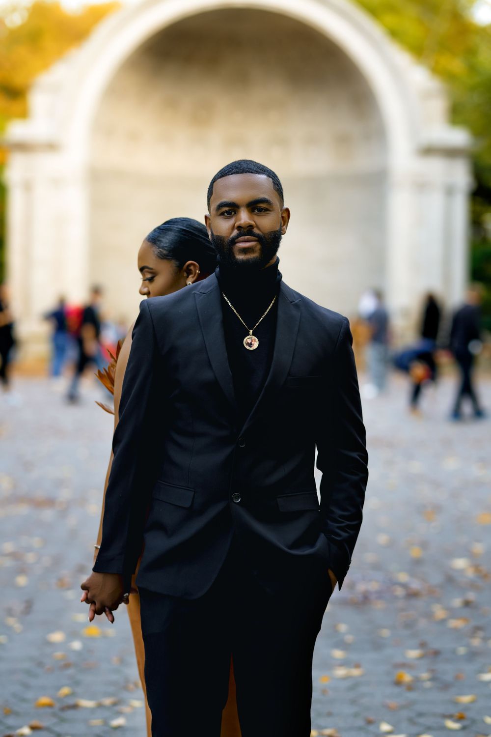 Stylish person wearing a black suit and turtleneck stands outdoors with autumn leaves scattered on the ground. Engagement photoshoot