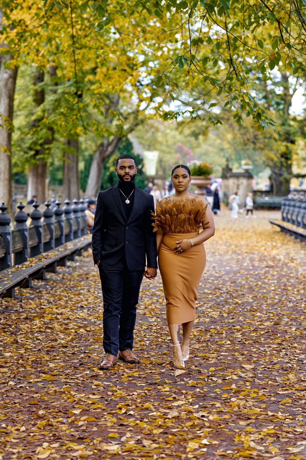 Stylish couple walks together on autumn path covered in yellow fallen leaves beside ornate metal railing. Engagement photoshoot central park