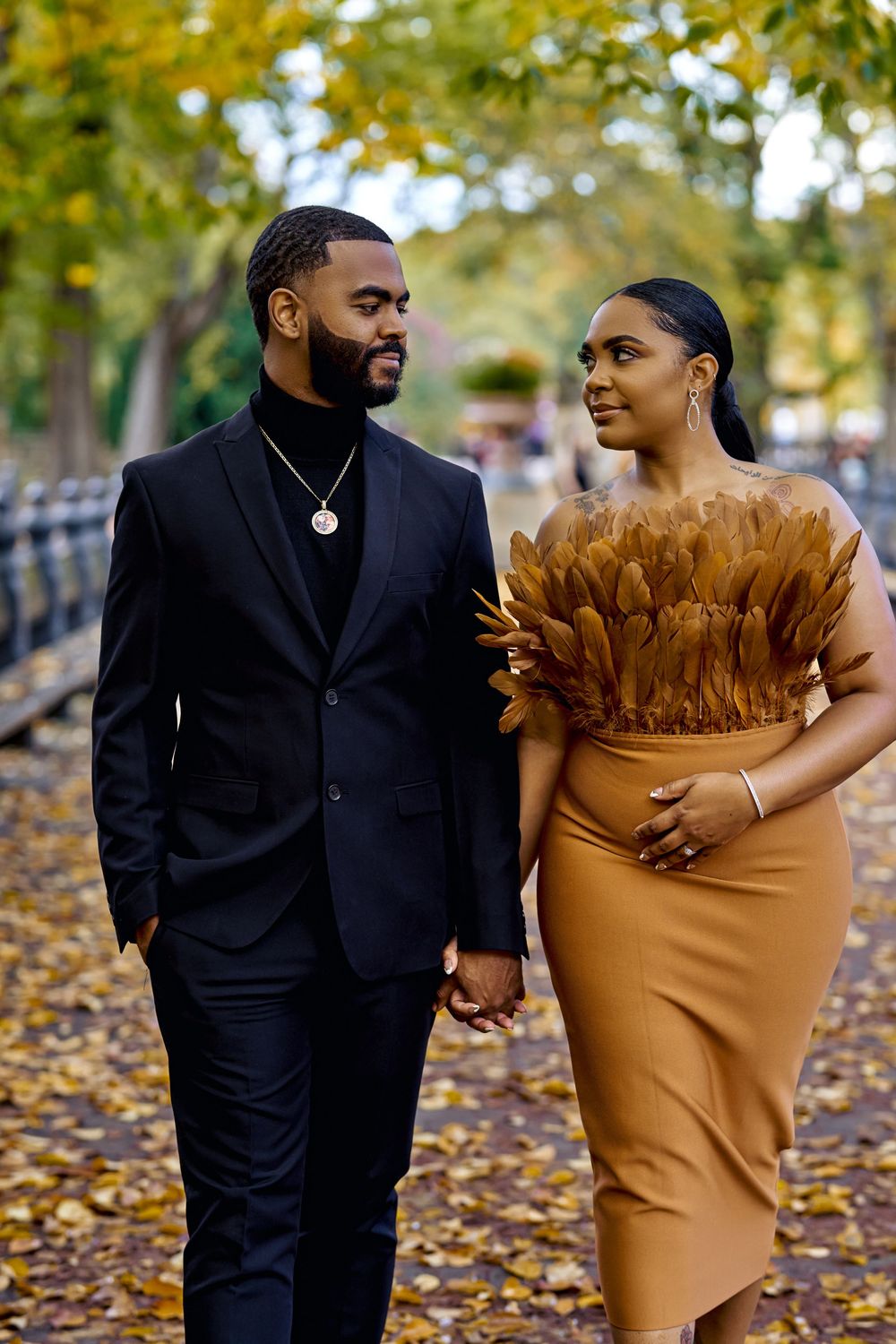A couple in formal attire walking along a leaf-covered pathway in autumn, with black suit and caramel dress details. Engagement photoshoot