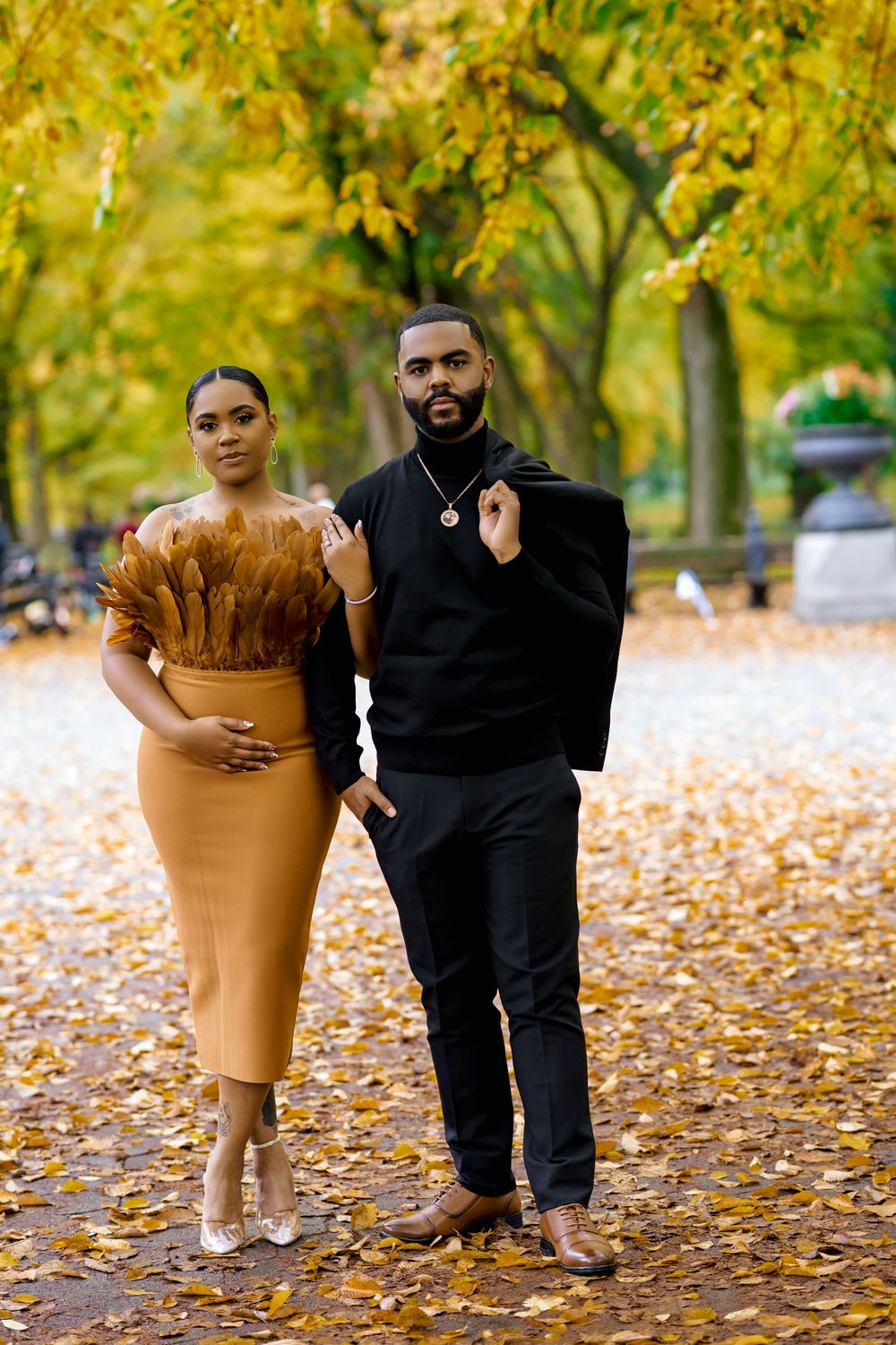 A couple poses together in a park during autumn with fallen leaves and golden trees in the background. Engagement photoshoot central park