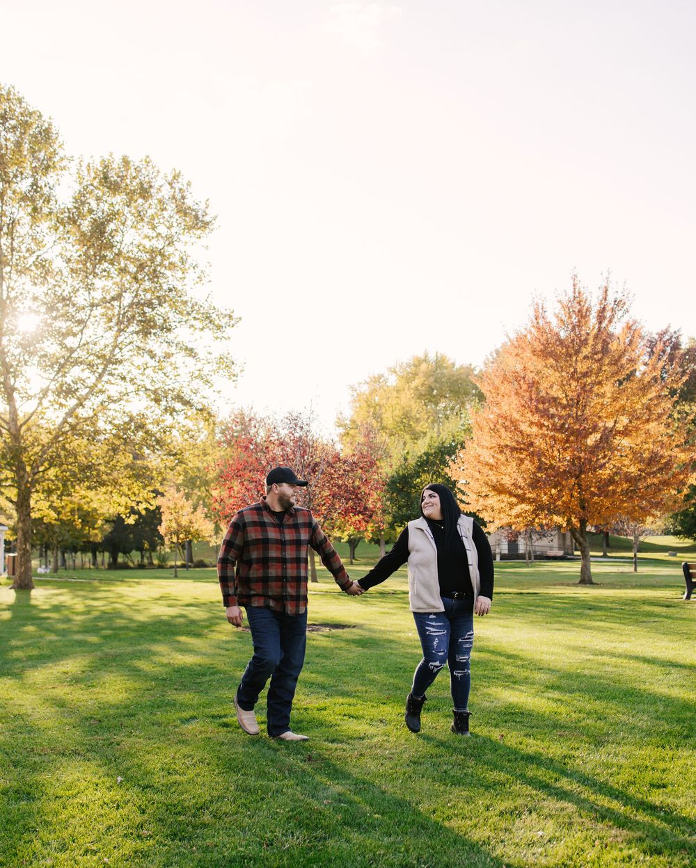 Couple holding hands walking through a lush green park with vibrant autumn foliage trees in golden sunlight.