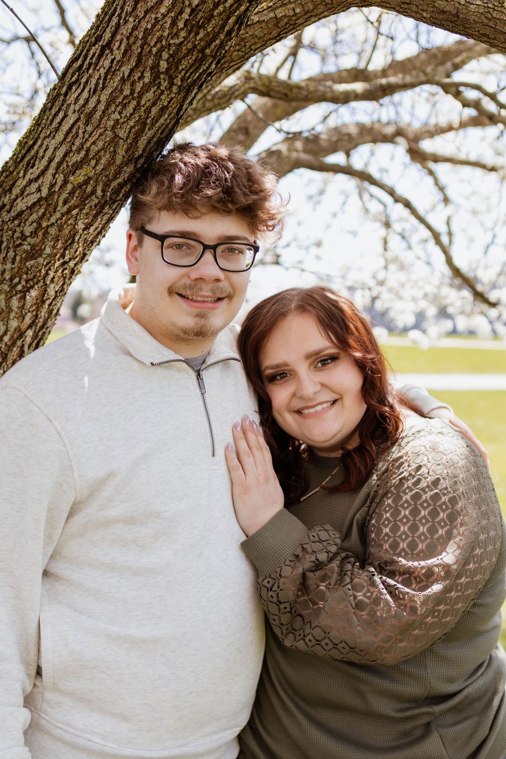 A couple poses together under a large tree branch on a bright spring day.