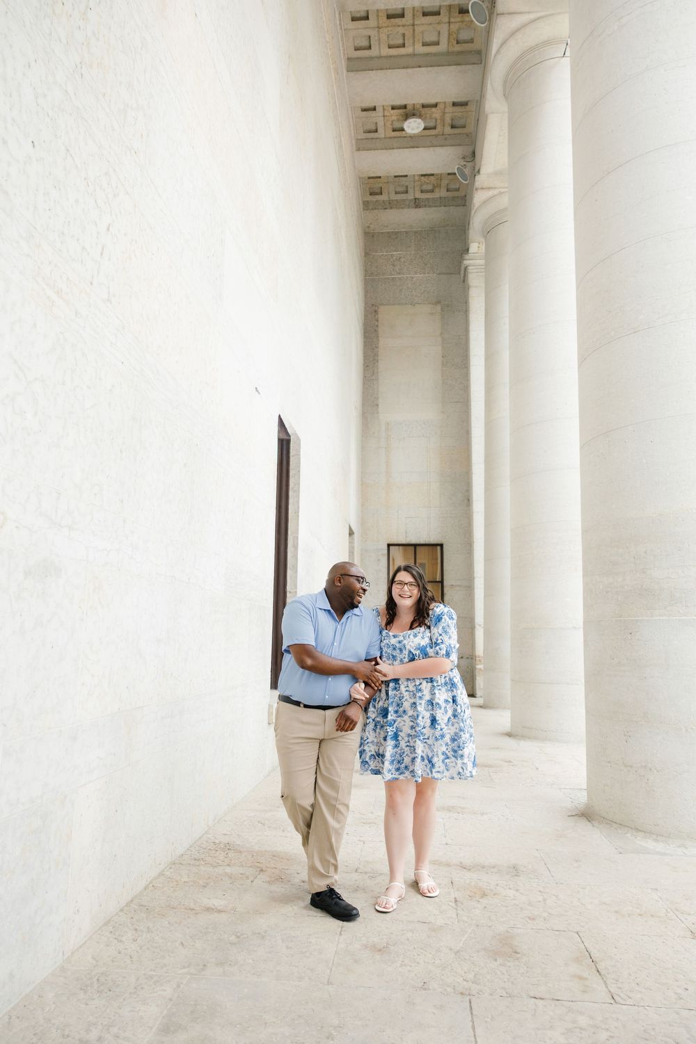 Couple walking hand in hand between grand white columns at a classical monument, woman in floral dress, man in blue shirt.