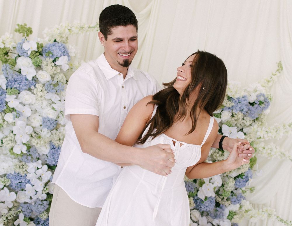 Happy couple dancing in white outfits in front of a blue and white hydrangea floral wall backdrop.
