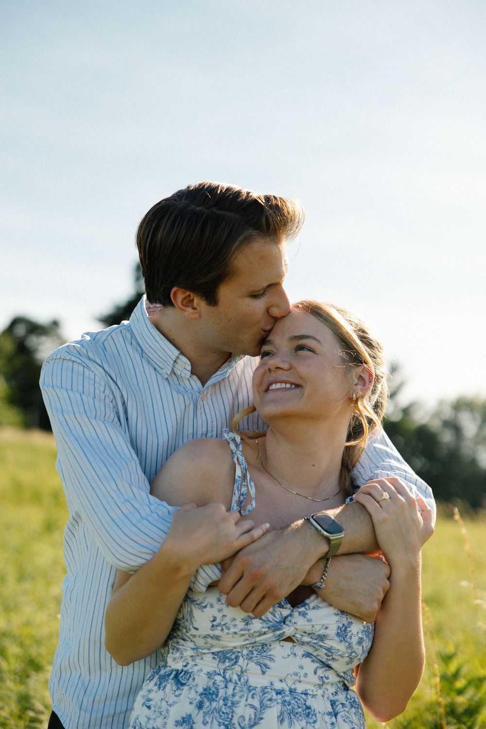A couple embraces in a sunny green field, man kissing womans forehead while she smiles joyfully.