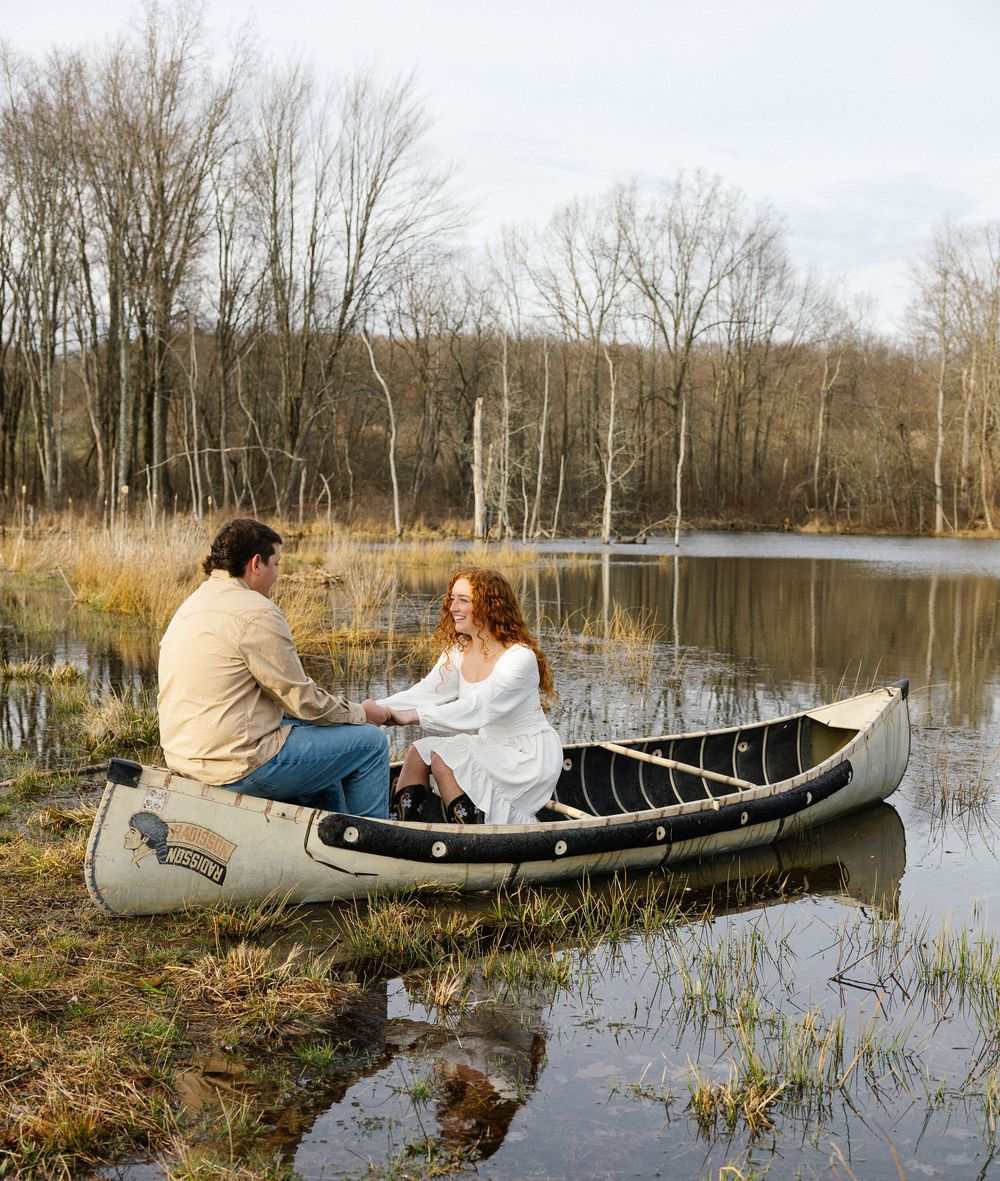 A couple sits facing each other in a canoe on a calm lake surrounded by bare trees and marsh grass.