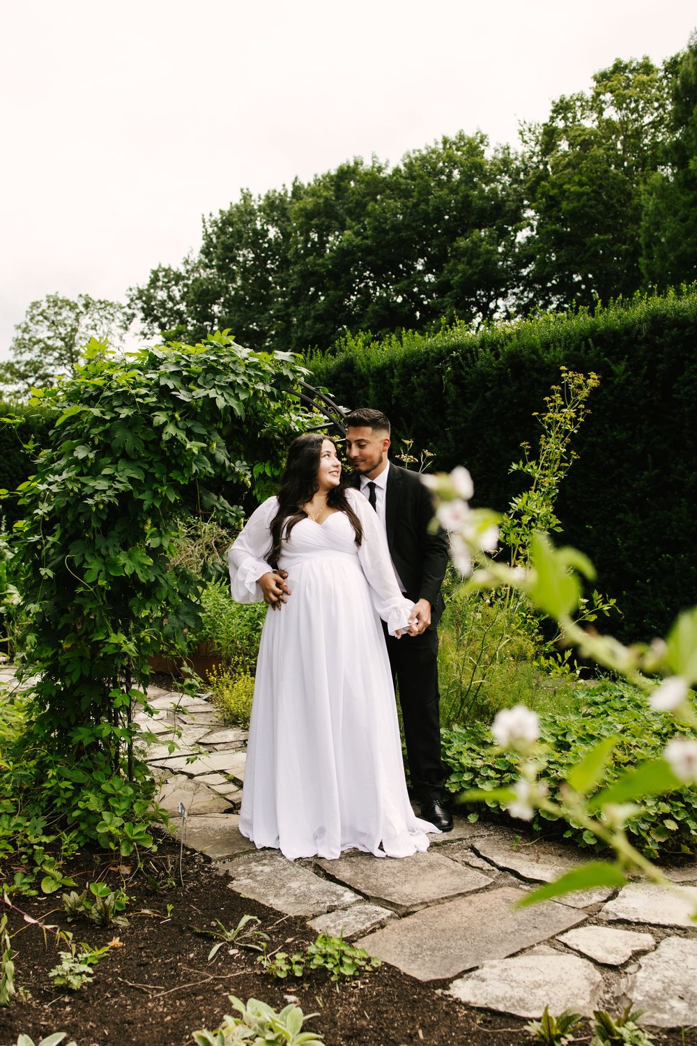 A couple in wedding attire poses together on a stone path surrounded by lush greenery in a garden setting.
