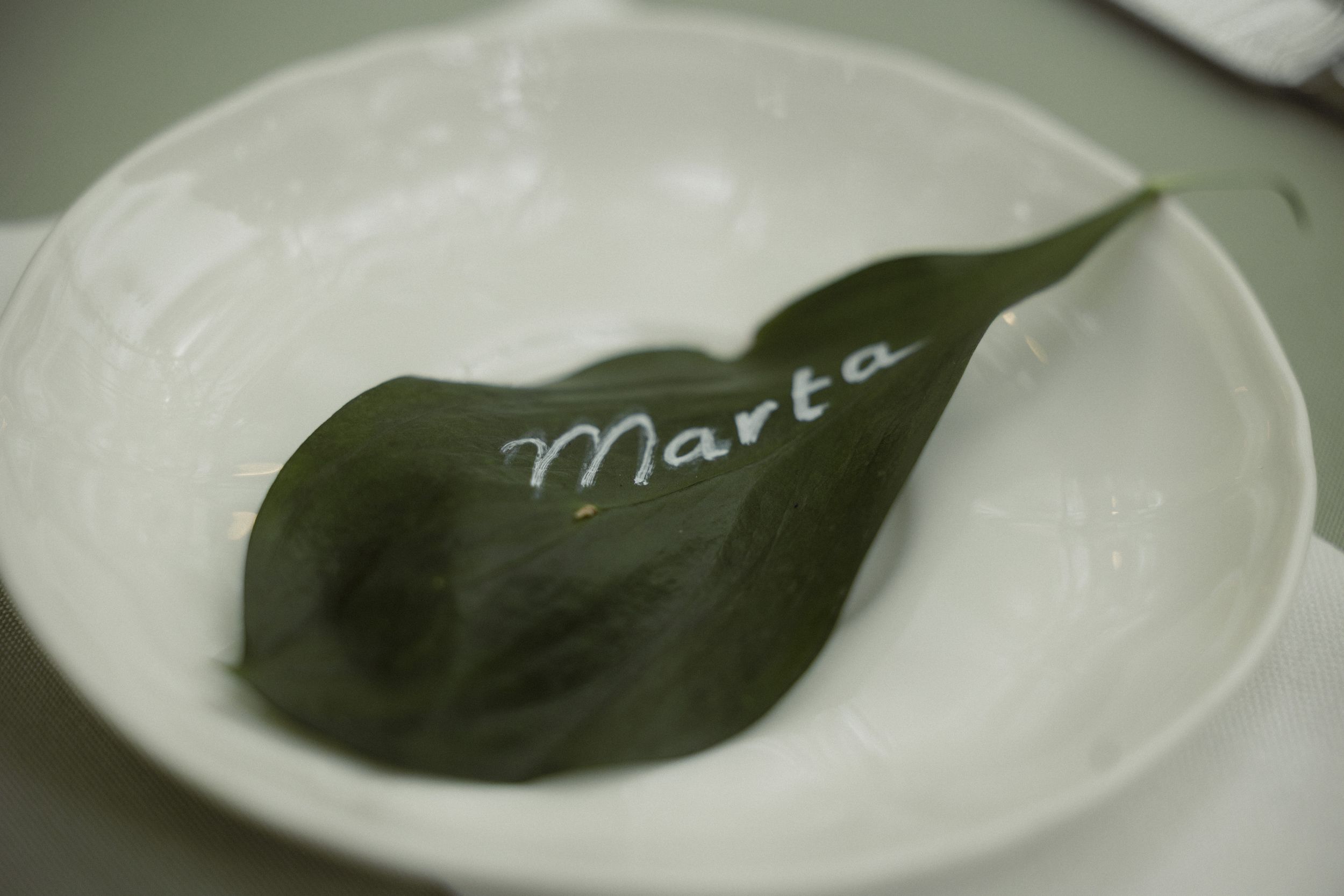 A green leaf place card with the name 'Marta' written in white chalk rests on a white plate.