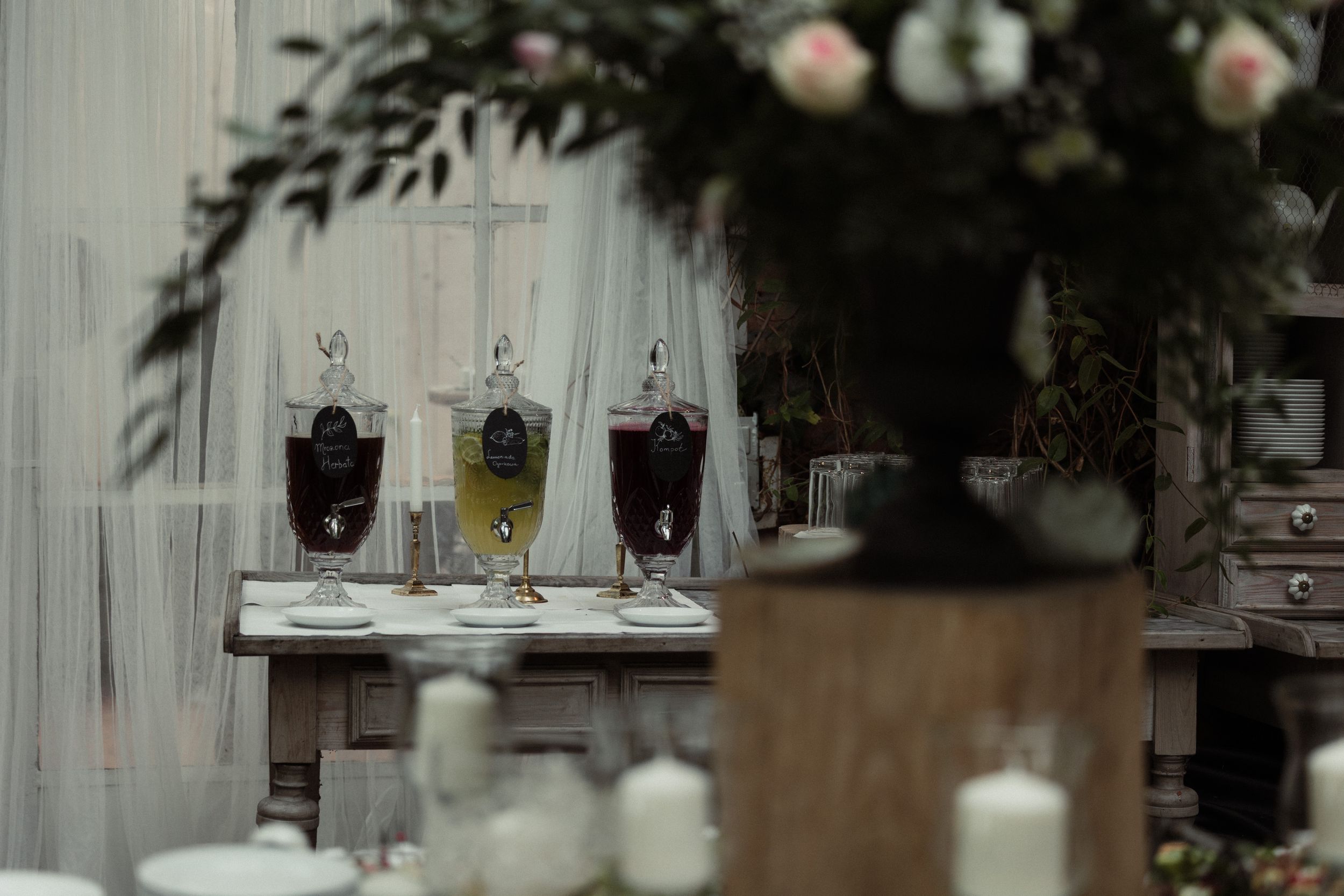 Three vintage-style beverage dispensers with different colored drinks sit beneath floral decorations at a rustic event setup.
