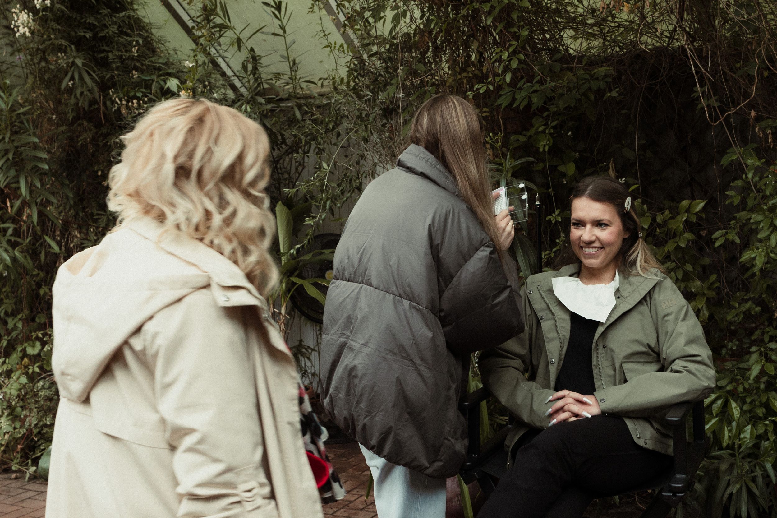 Three people in winter coats stand together having a casual conversation outdoors near green foliage.