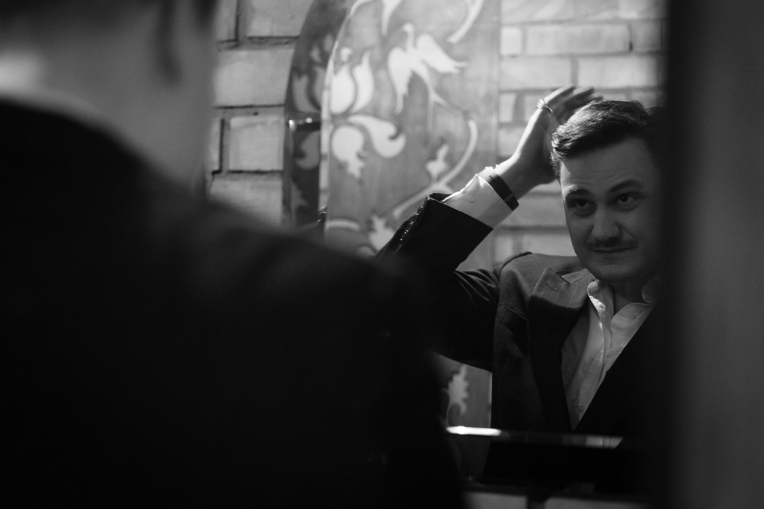 A black and white portrait photo of someone sitting indoors by a brick wall with atmospheric lighting.