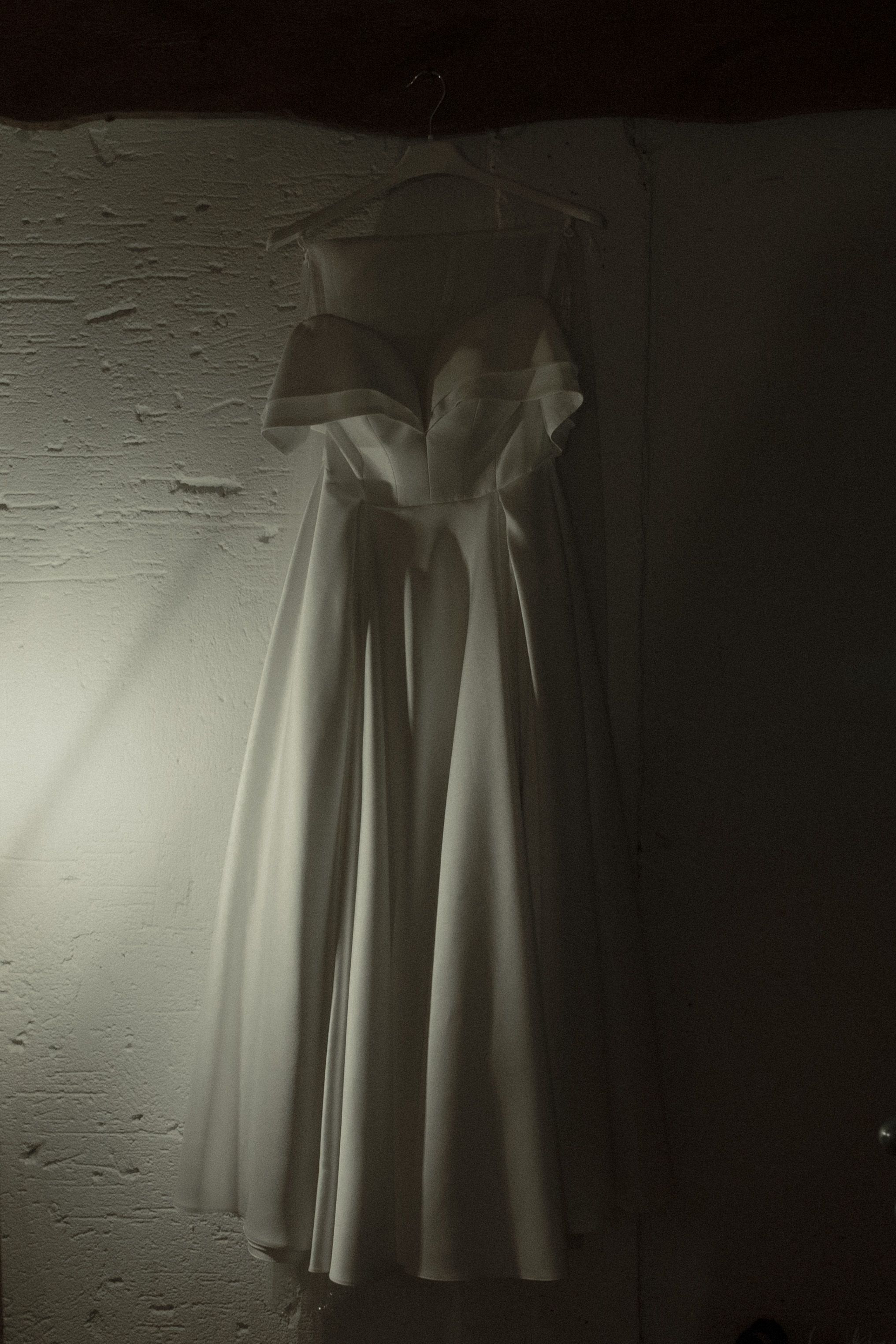 A white vintage wedding dress hanging on display in moody dramatic lighting against a textured wall.