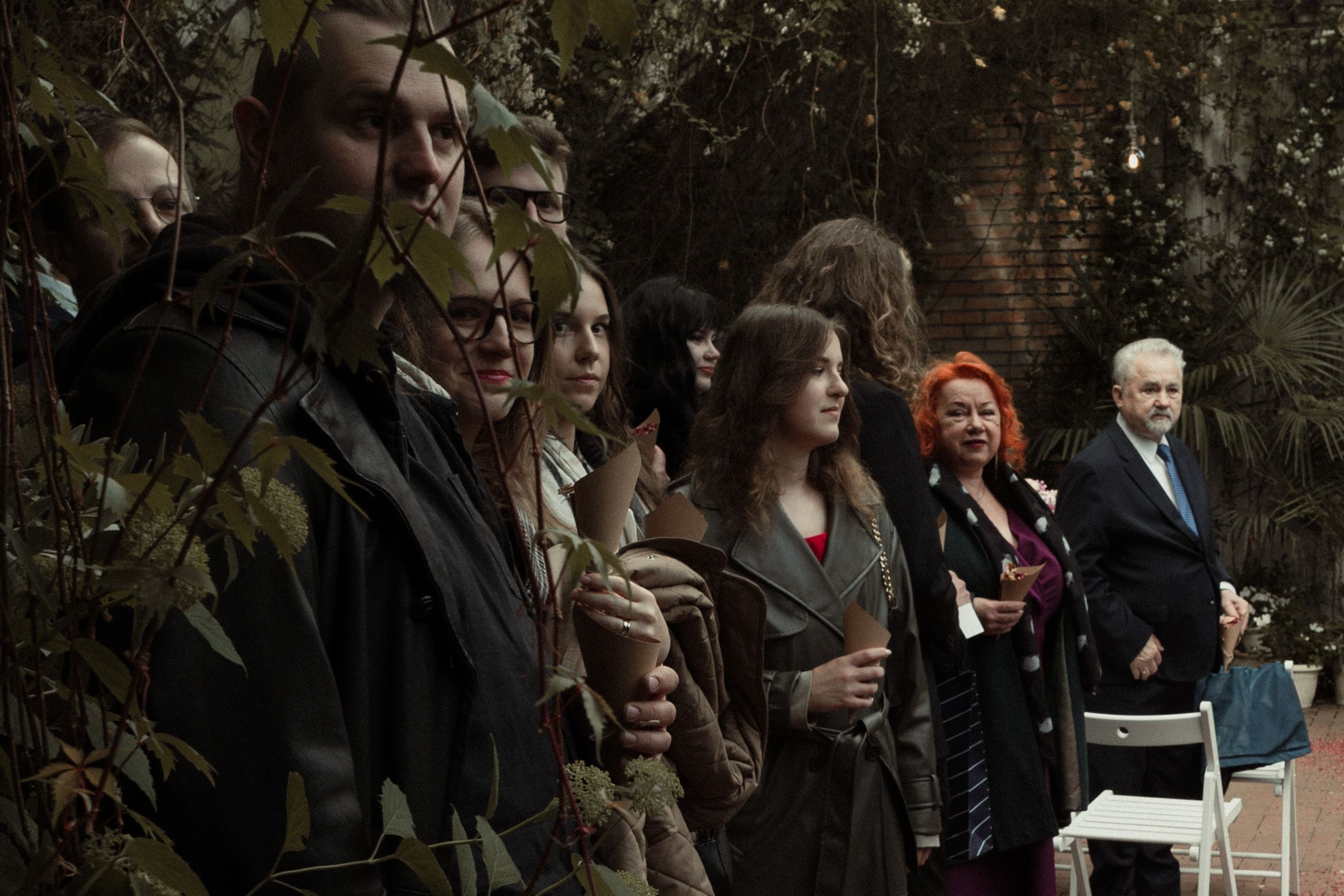 A series of solemn attendees gather outdoors for what appears to be a funeral or memorial service.