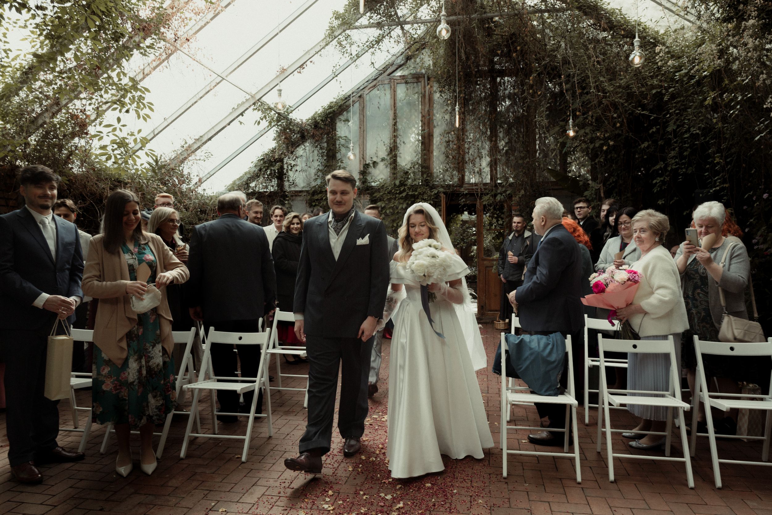 An outdoor wedding ceremony moment captured beneath trees with white folding chairs and guests gathered.
