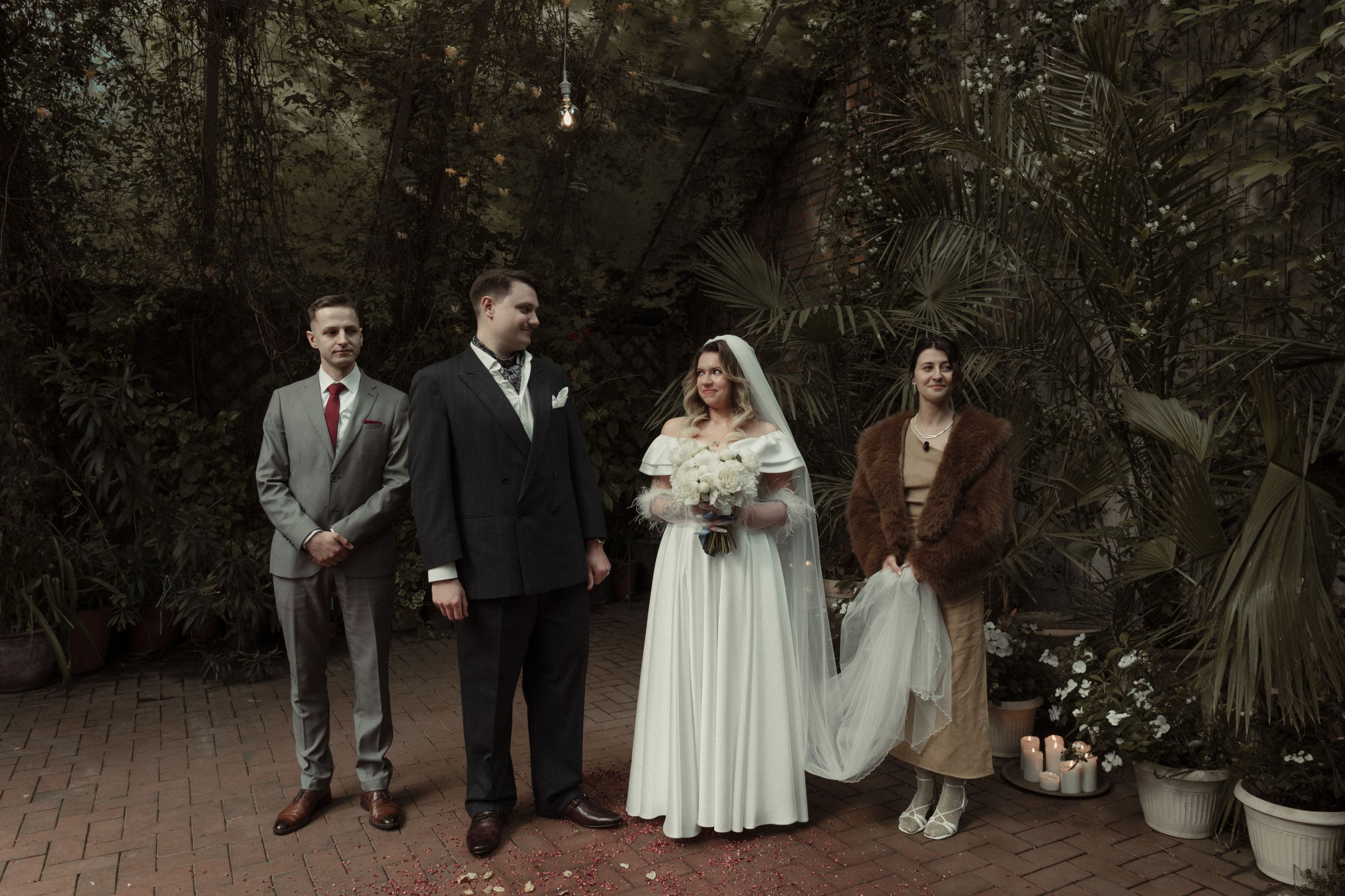 A group of four well-dressed wedding guests in vintage attire stand together on a brick patio surrounded by greenery.