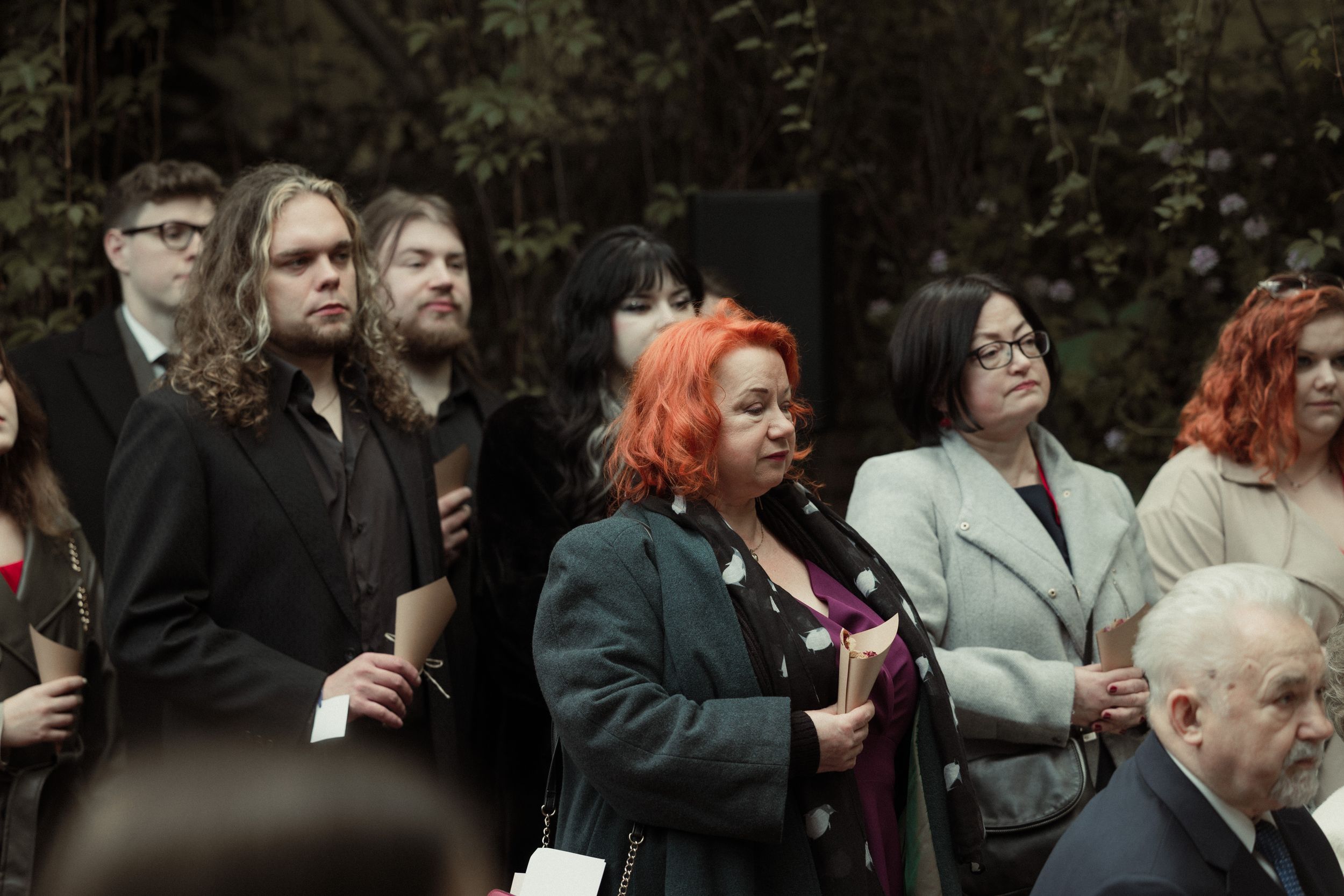 A group of guests stand attentively outdoors during an autumn gathering with dark foliage in the background.