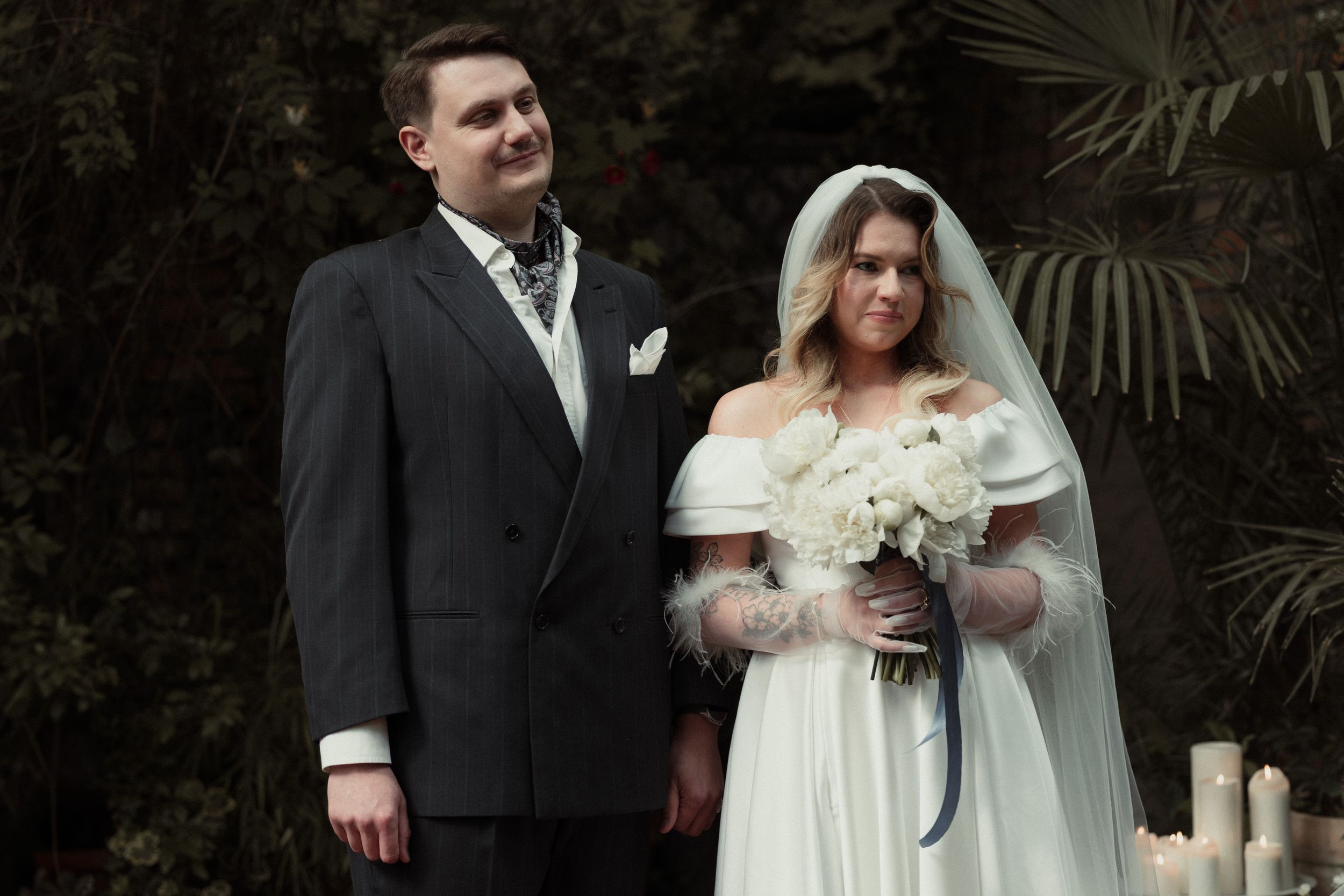 A bride in winter white dress with fur stole and groom in dark suit pose for their wedding photo against greenery.