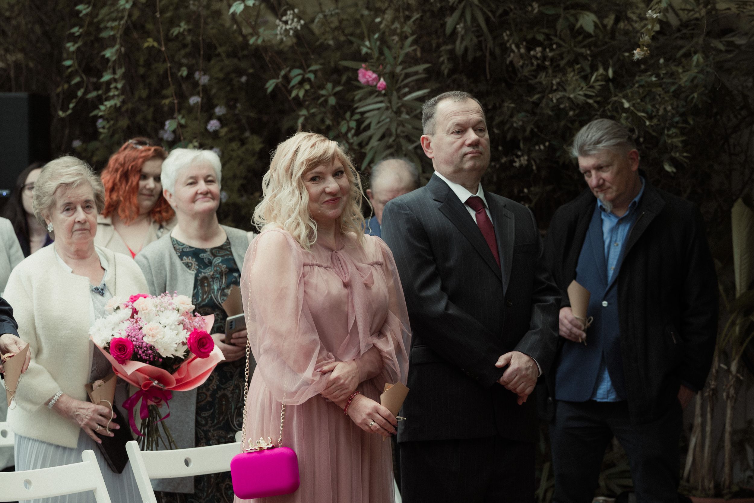 People gather outdoors for a formal event, with attendees wearing dressy attire in shades of pink and black.