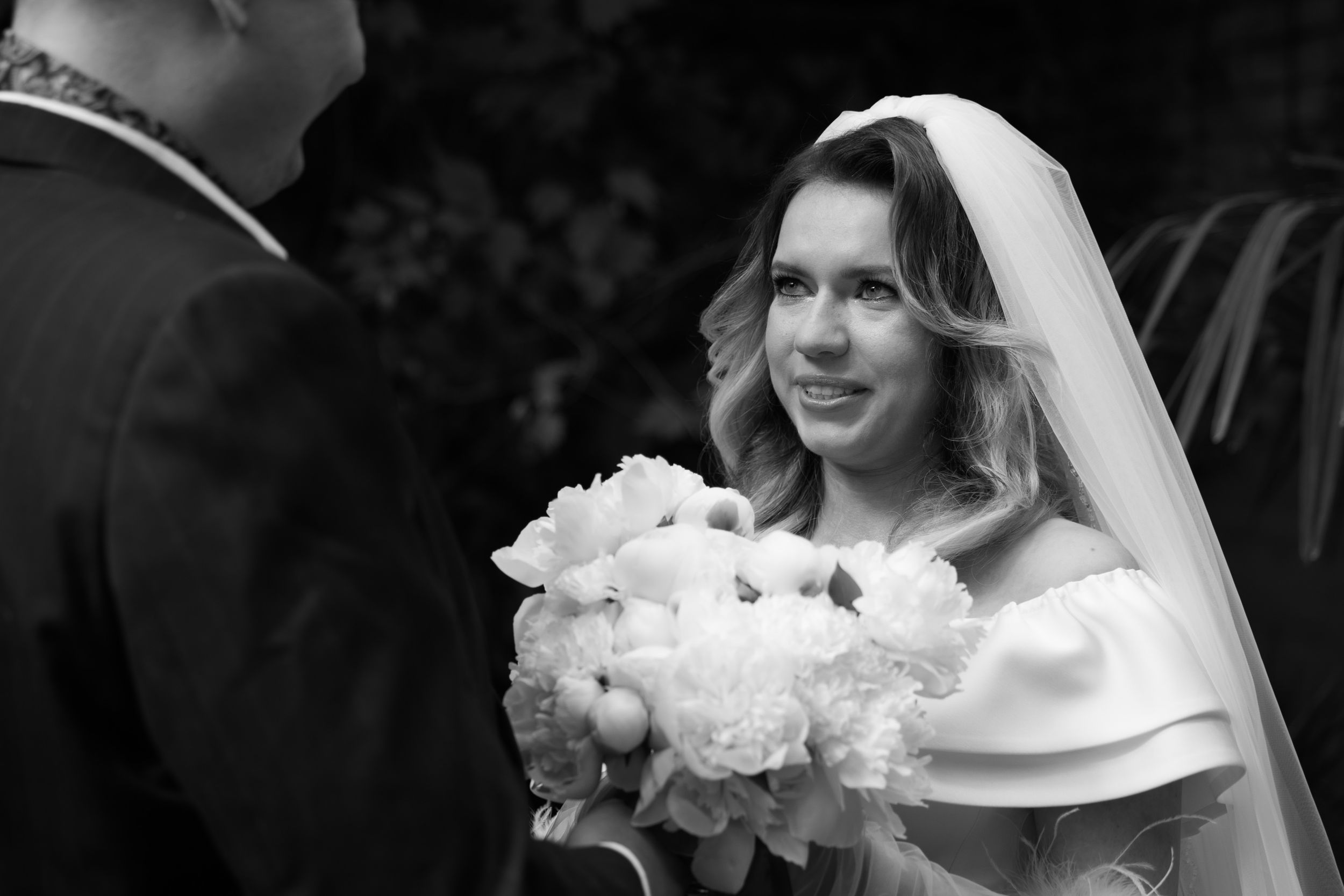 A bride in a white gown holds a bouquet of white peonies while wearing a traditional veil during a wedding ceremony.