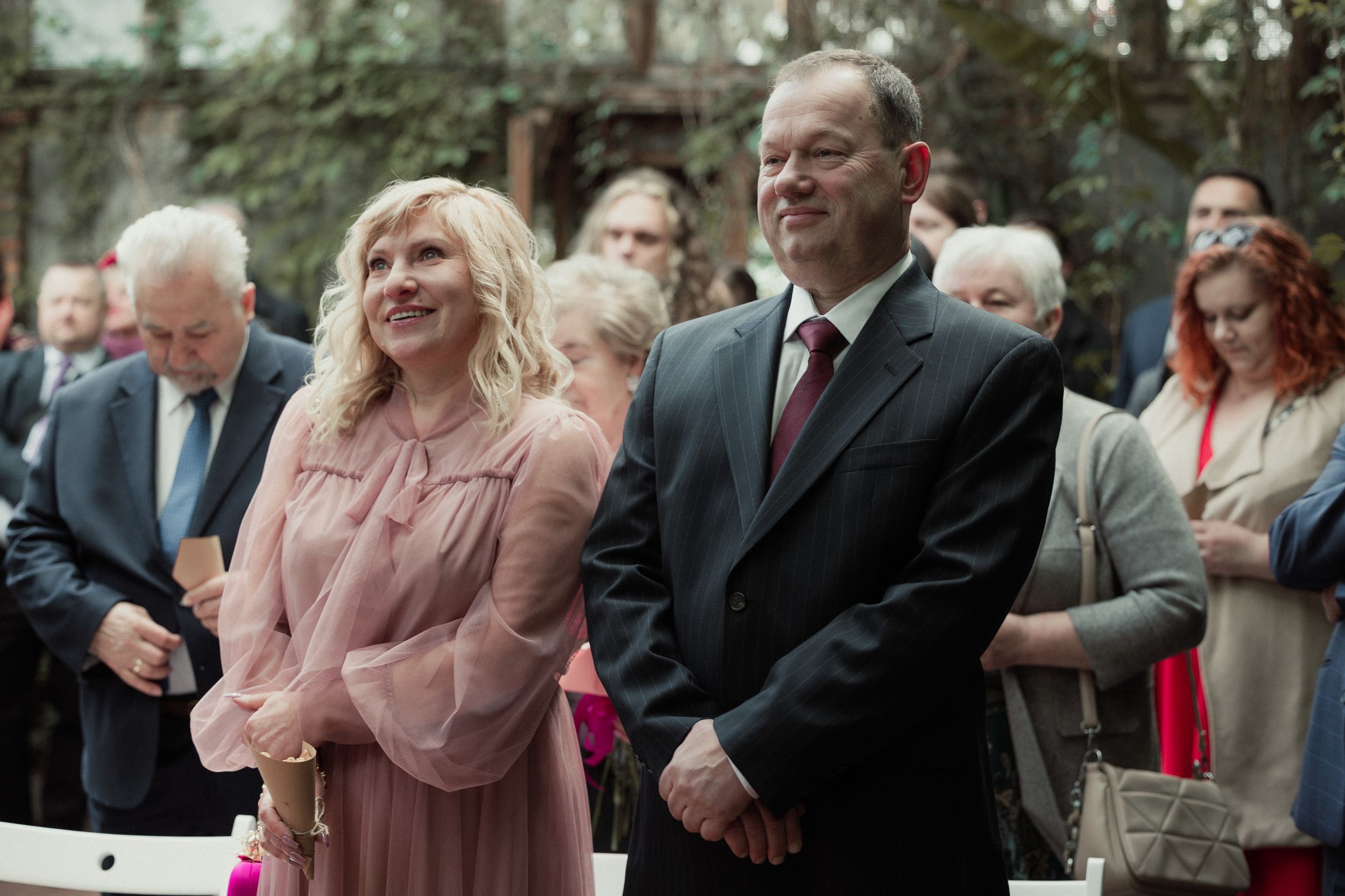 Two guests at a formal outdoor event stand together, with the woman wearing a pink blouse and the man in a dark suit.