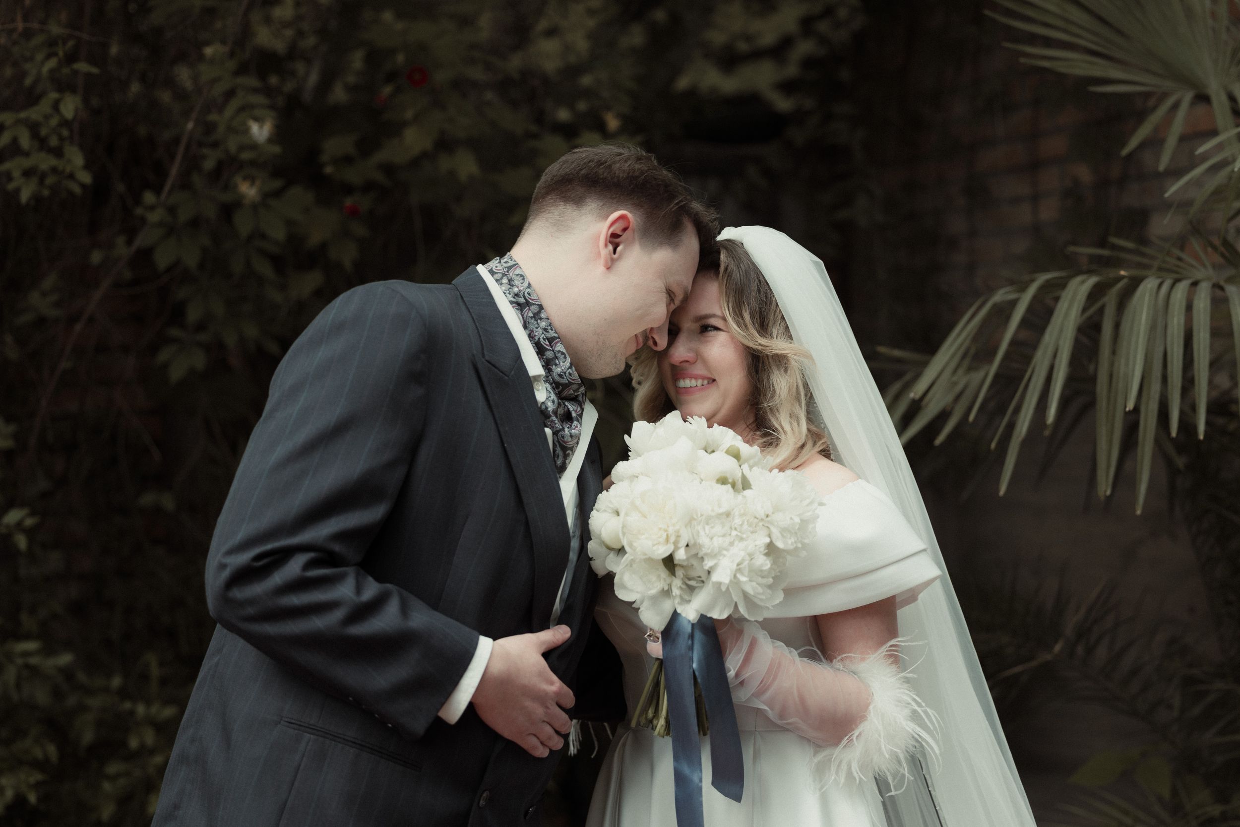 A tender moment shared between a couple at their winter wedding with white floral bouquet and grey suit against greenery.