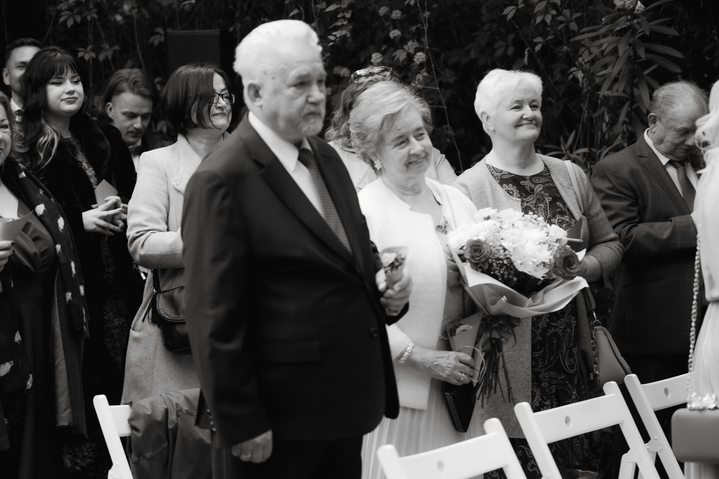A black and white wedding ceremony photo showing guests and family members gathered outdoors with bouquets.