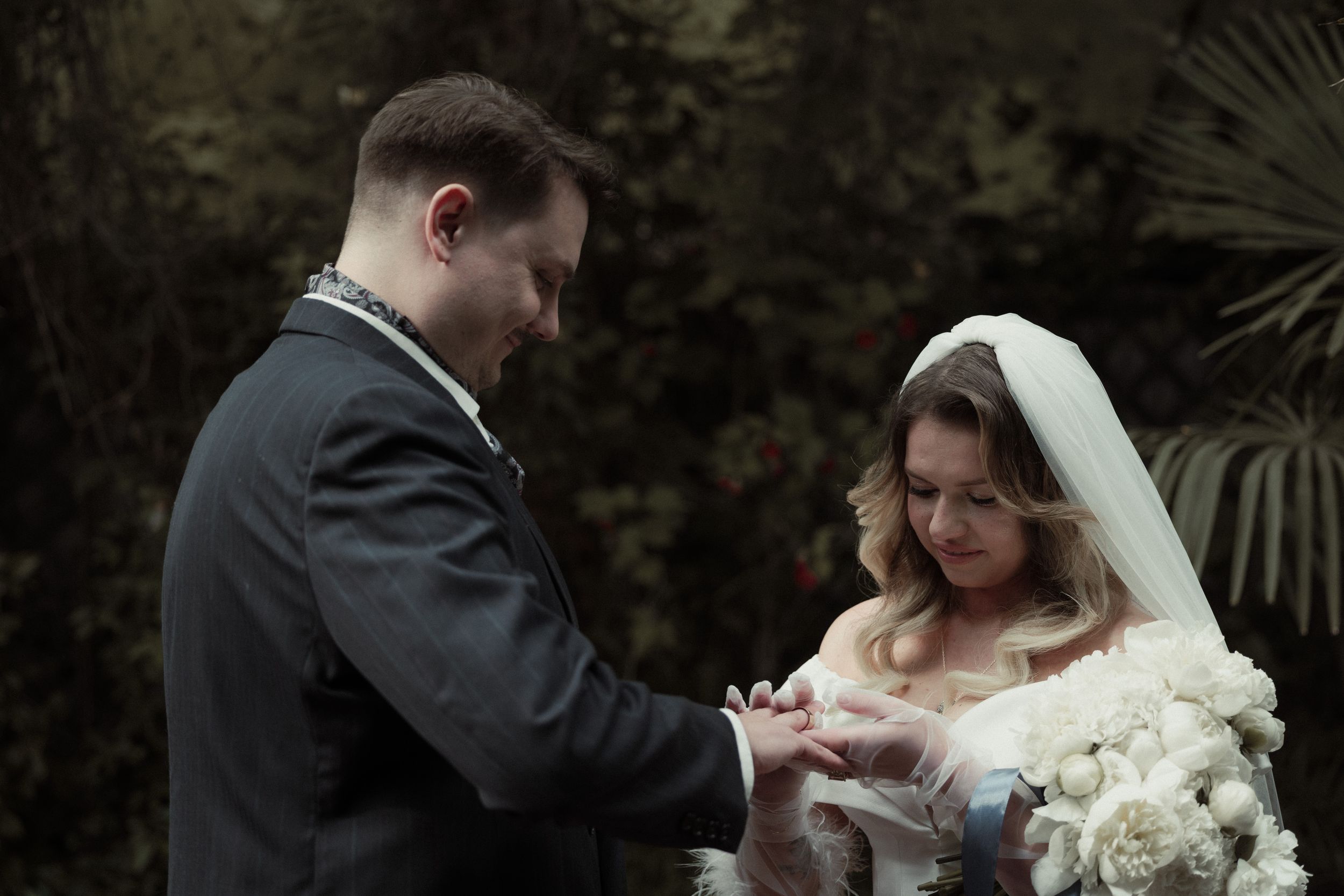 A couple exchanges rings during a wedding ceremony in a garden setting with dark foliage in the background.
