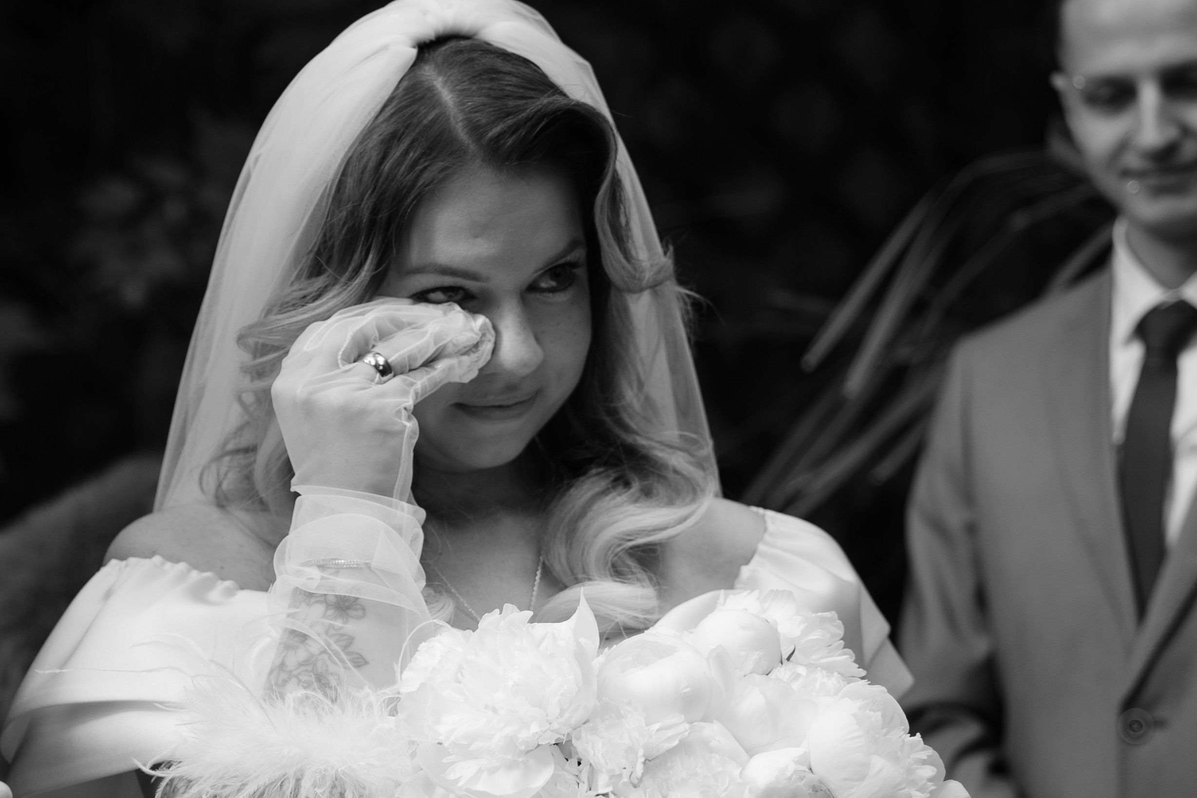 A happy bride in a white veil and dress wipes a joyful tear from her eye during a black and white wedding moment.