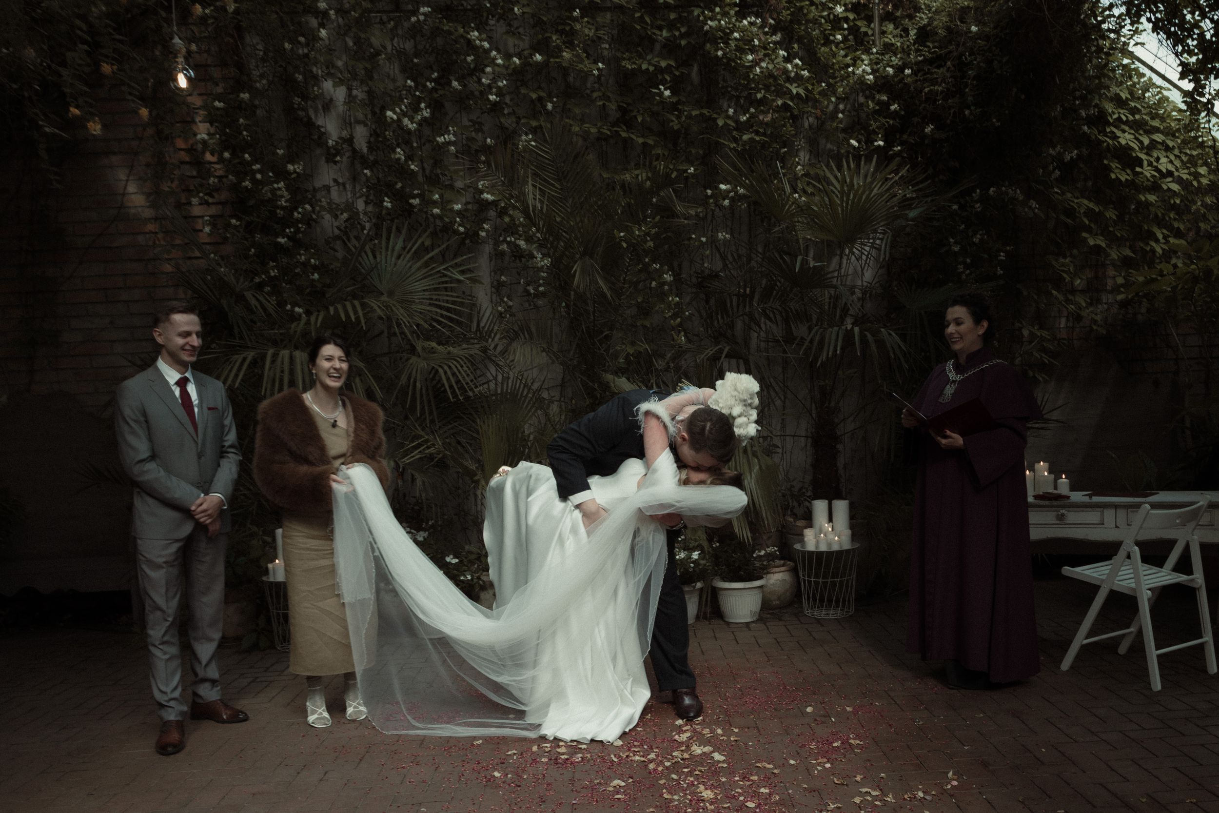 People gather together for an intimate outdoor wedding ceremony as rose petals scatter in a romantic garden setting.