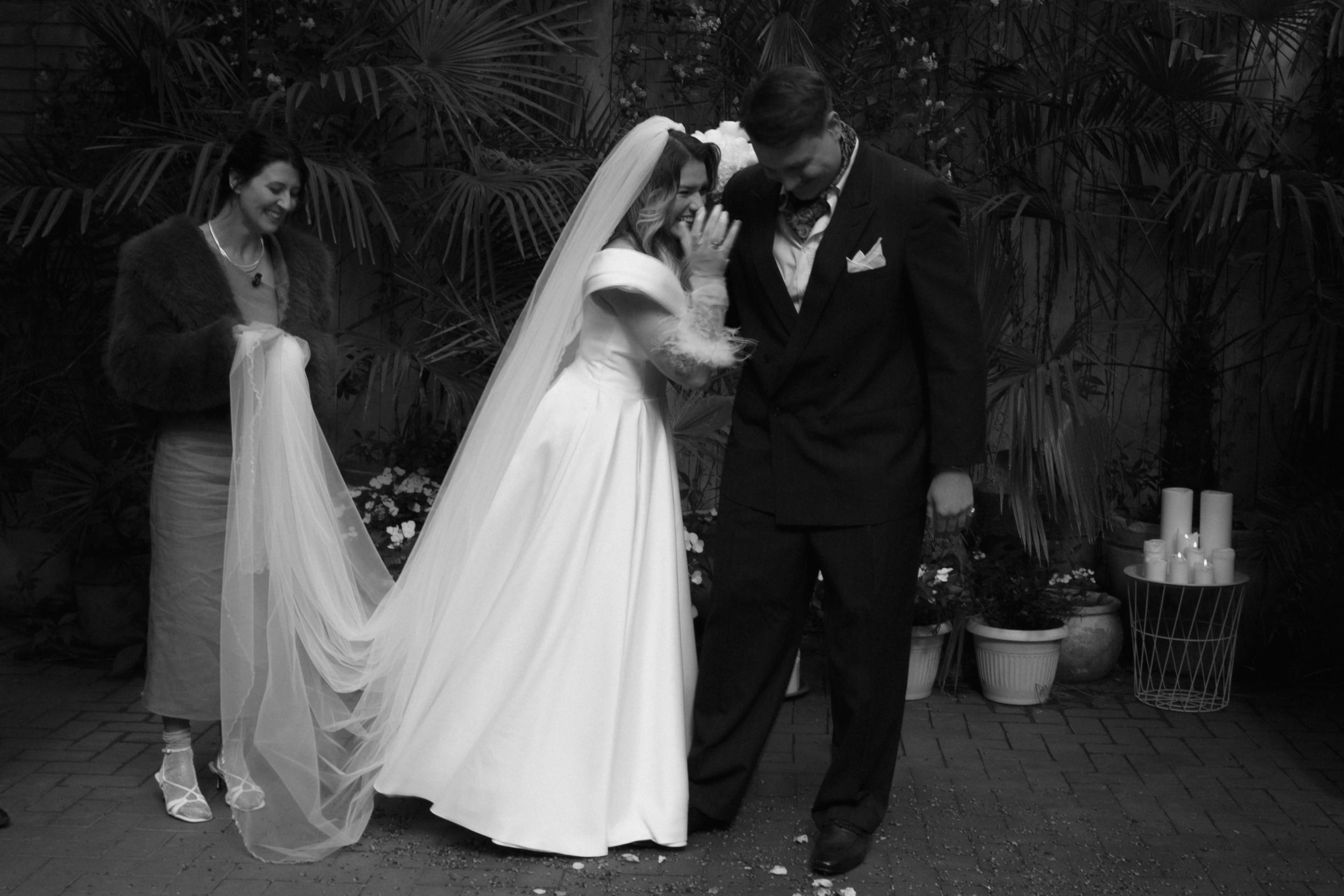 A black and white photo captures a tender moment at a wedding ceremony with a bride in white and guests in formal attire.