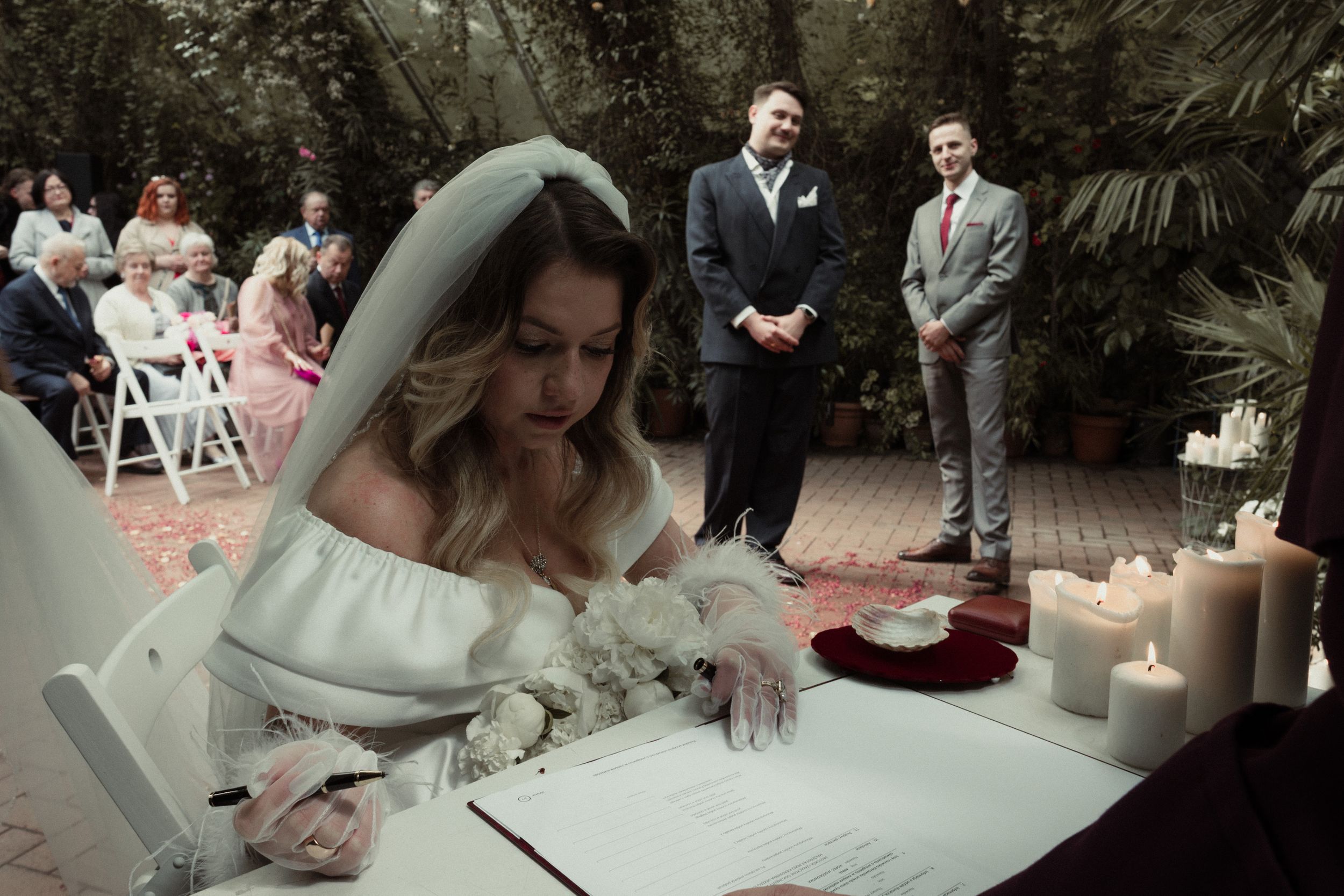 Wedding ceremony moment as someone signs documents at outdoor garden venue with guests and white candles.