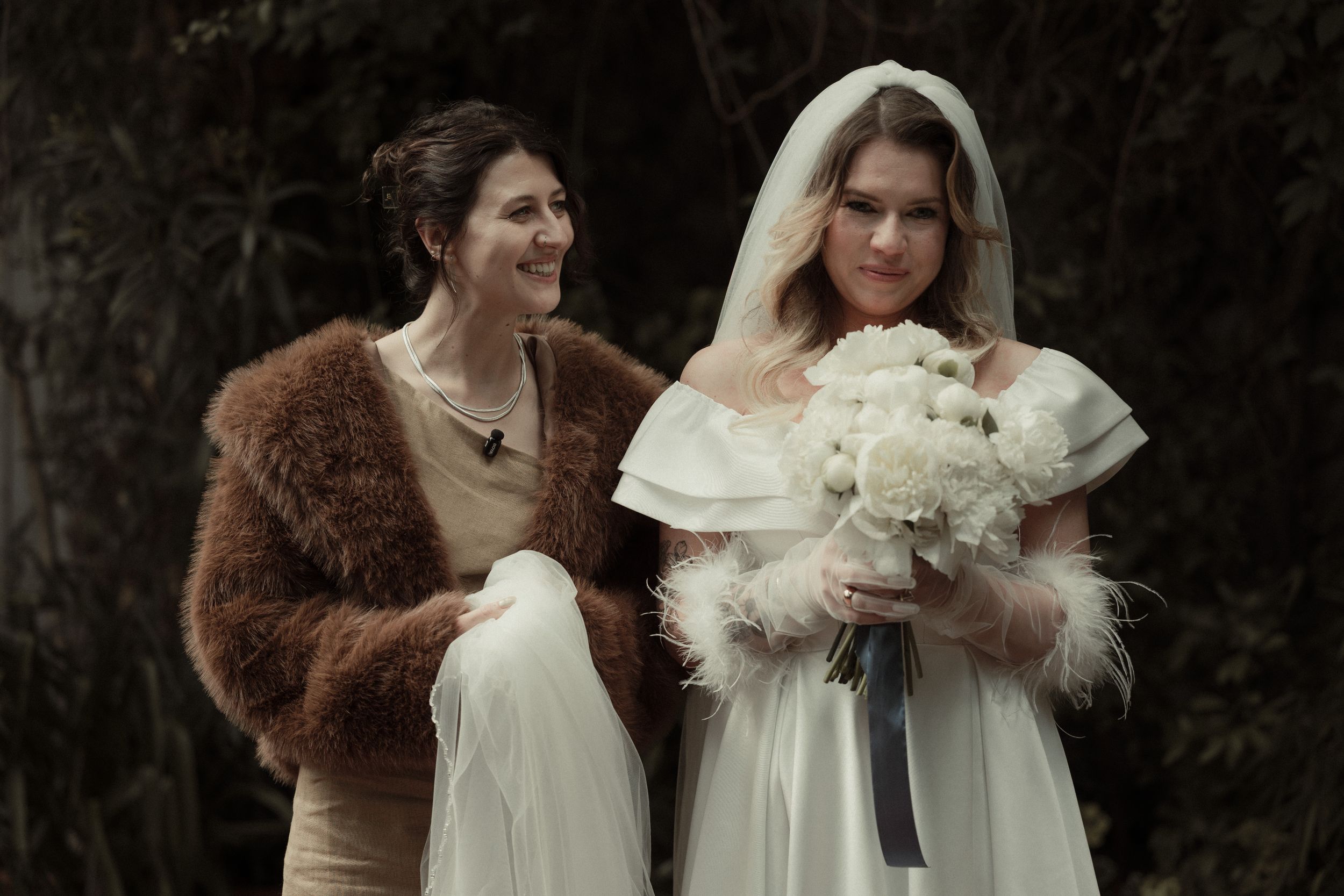 Two women in vintage winter wedding attire pose outdoors, one wearing a fur stole and the other in a white gown with veil.