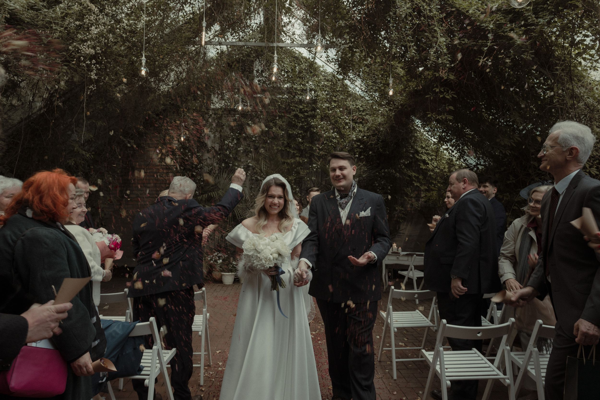 A couple walks down the aisle at an outdoor garden wedding ceremony while guests throw white confetti in celebration.
